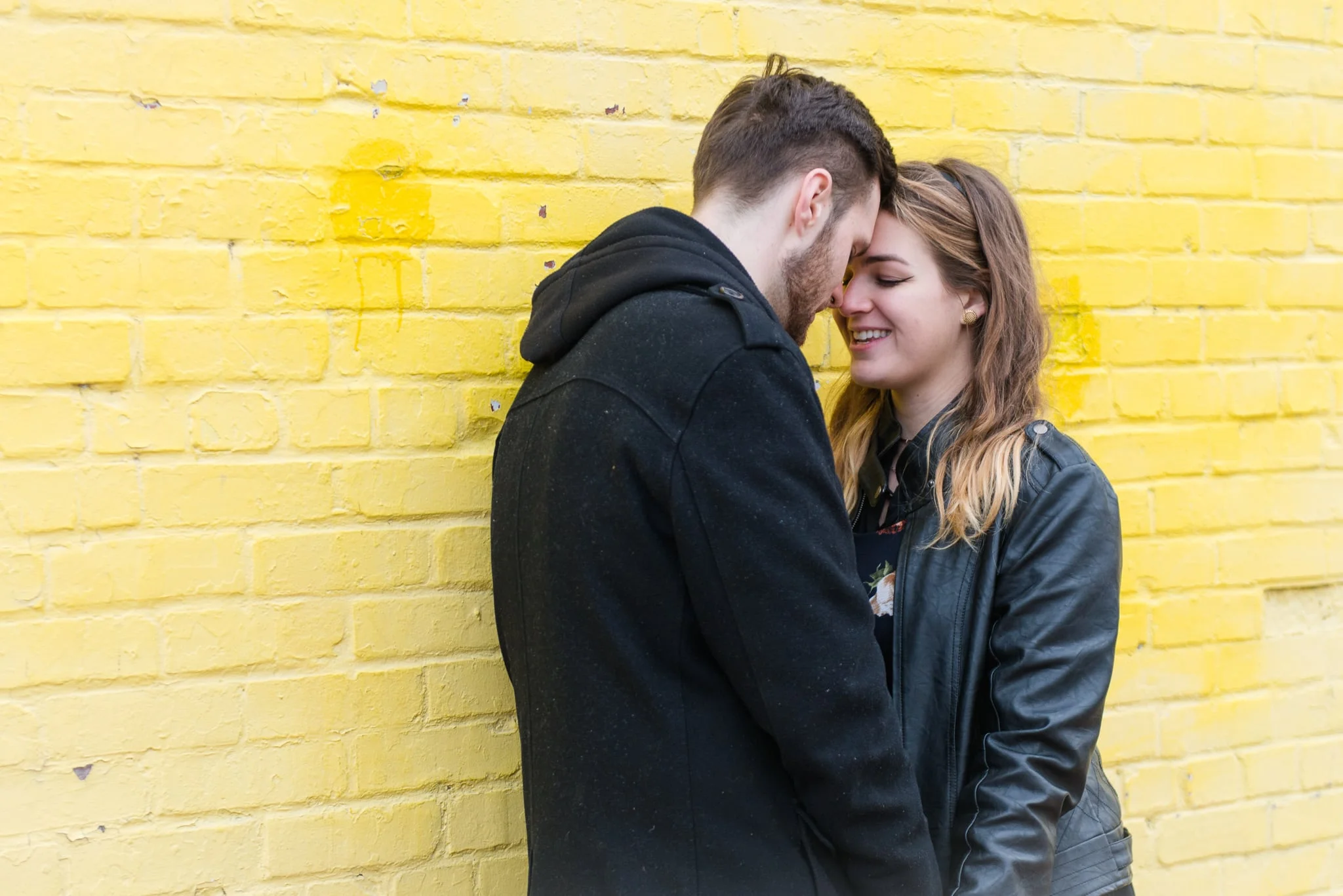 A young couple holds hands and touch foreheads together in front of a bright yellow brick wall during their portrait session in Old Town Alexandria
