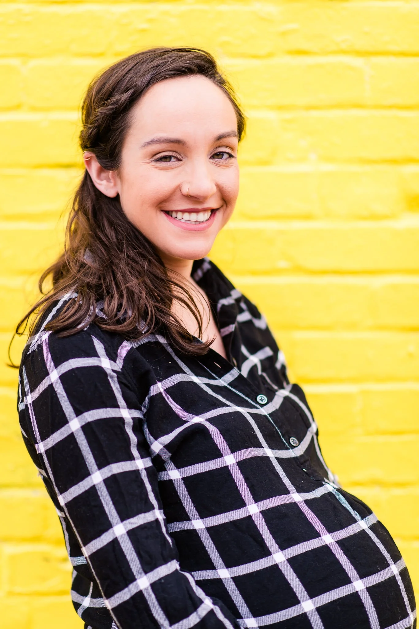 A pregnant woman poses in front of a bright yellow brick wall during a maternity session in Old Town Alexandria