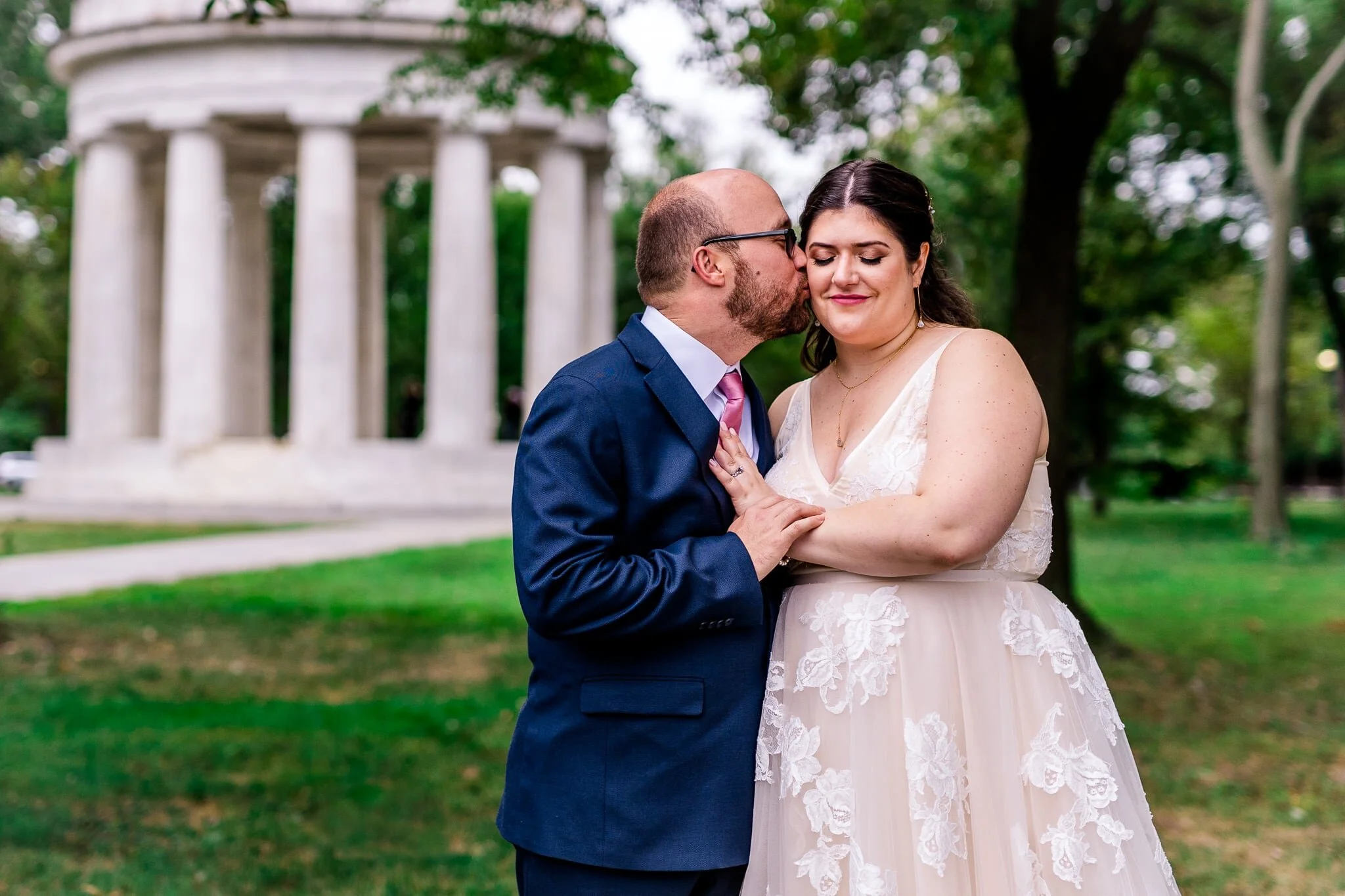 A groom kisses his bride on the cheek on their fall wedding day in front of the DC War Memorial in Washington, DC
