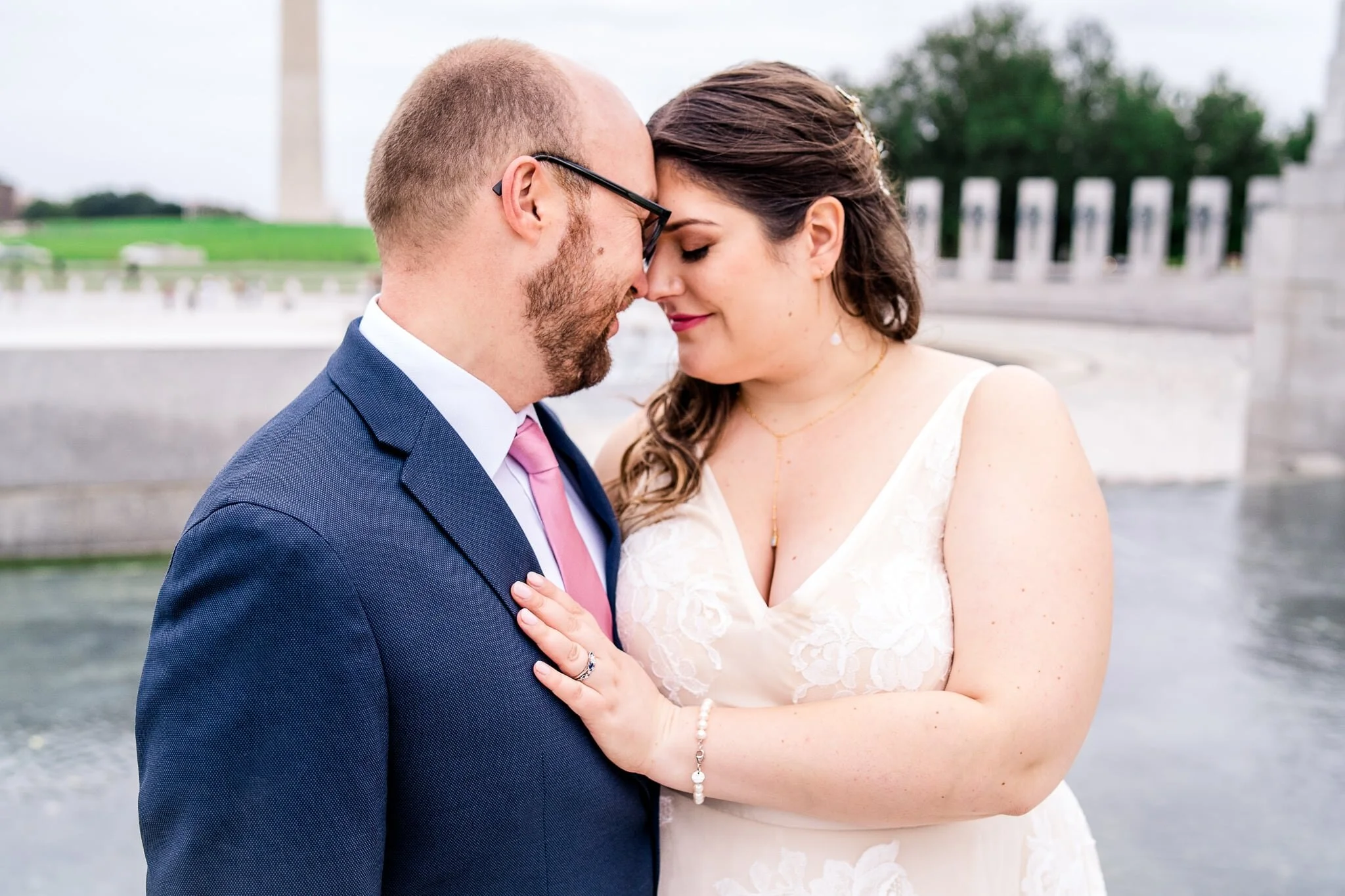 A bride and groom stand together with foreheads together and her hand on his chest on their fall wedding day at the WWII Memorial in front of the Washington Monument in Washington, DC