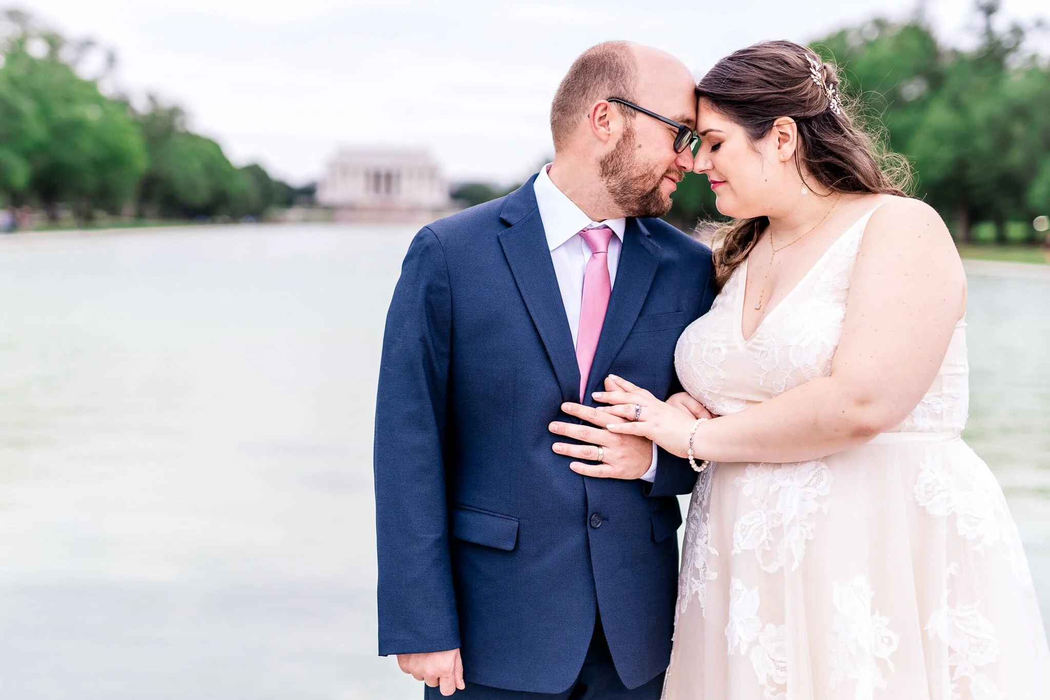 A bride and groom pose together arm-in-arm with foreheads and noses together on their fall wedding day at the Reflecting Pool in front of the Lincoln Memorial in Washington, DC
