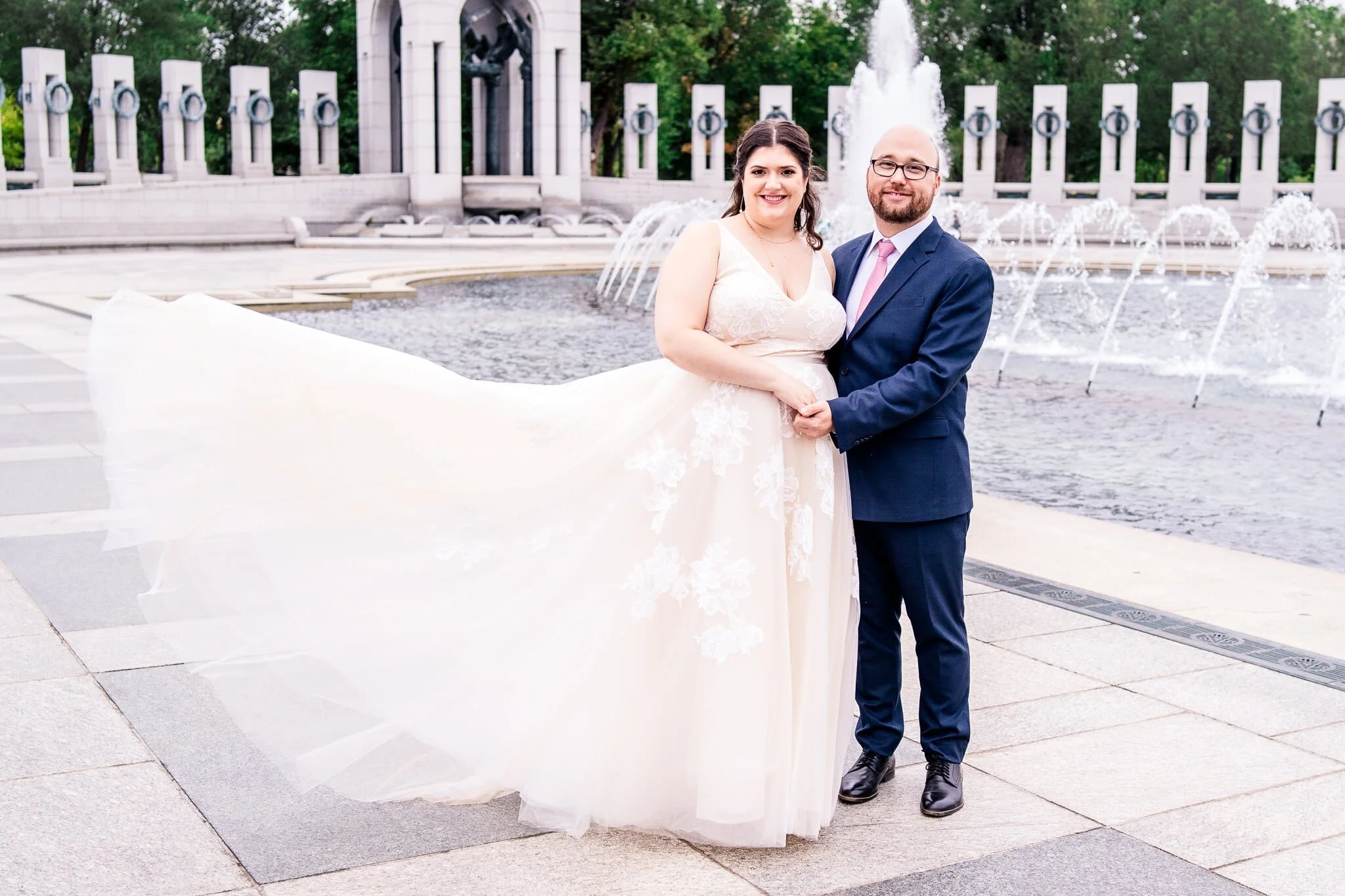 A bride and groom stand together as the bride’s dress dramatically blows in the breeze on their fall wedding day at the WWII Memorial in Washington, DC