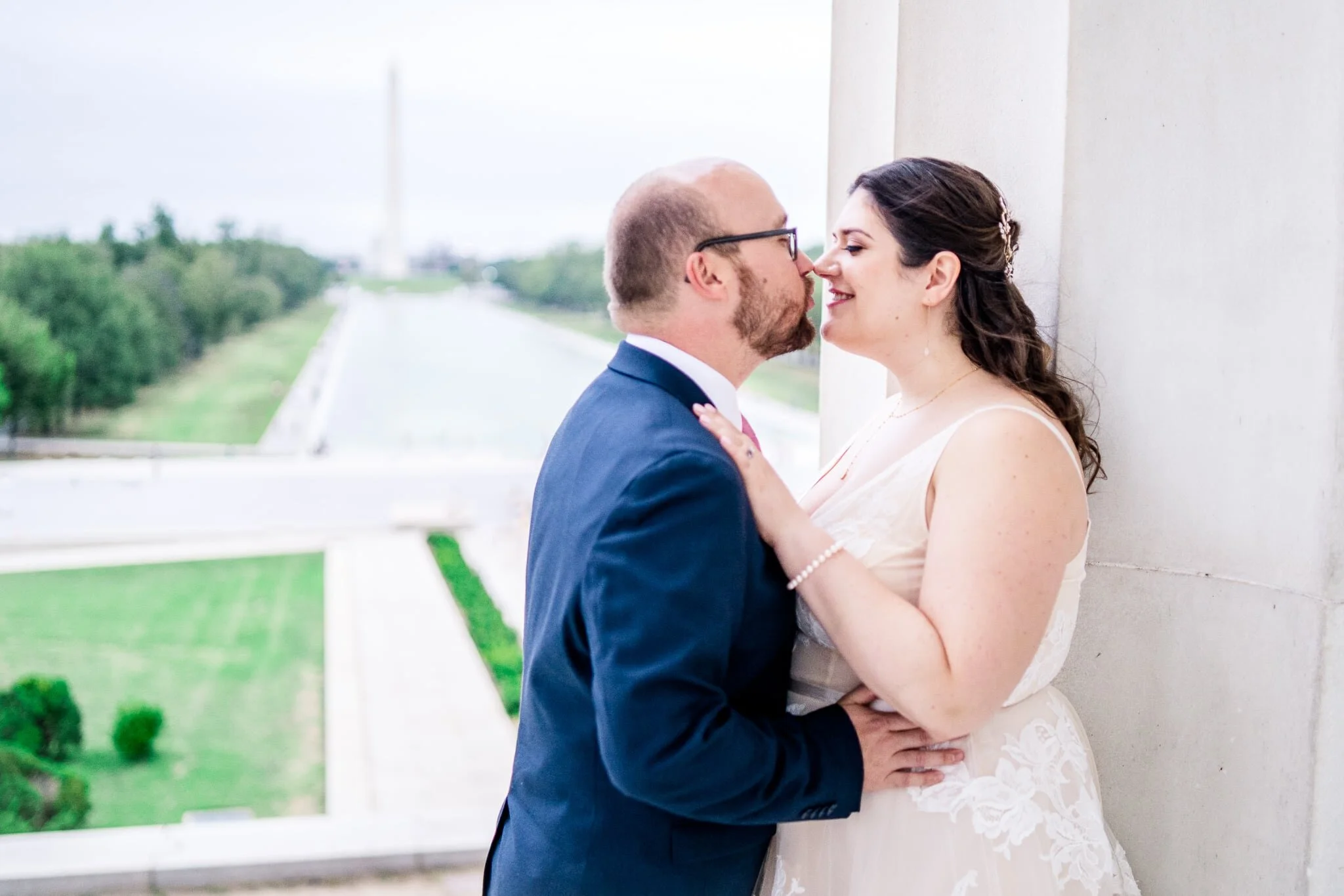 A bride and groom snuggle in together beside a marble column on their wedding day with a view of the reflecting pool and Washington Monument at the Lincoln Memorial in Washington, DC