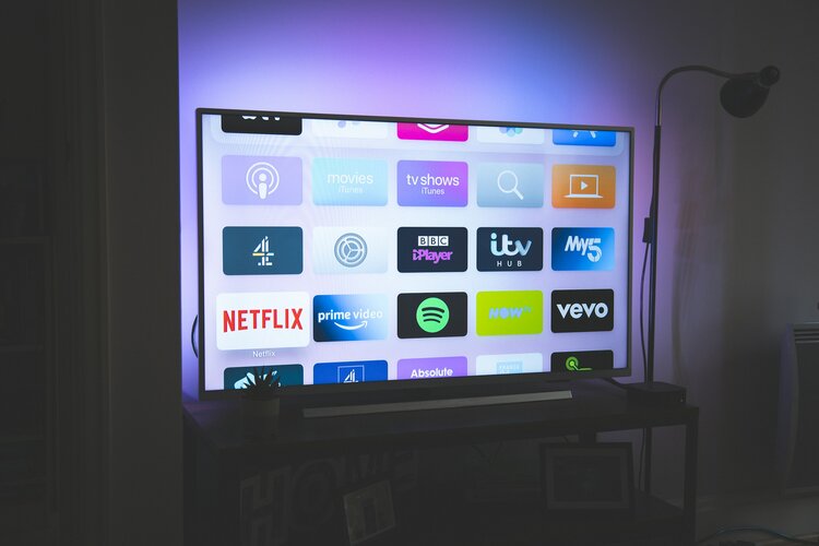 A smart TV on a stand. The tv displays icons from various tv applications (netflix, vevo, etc.) sitting in front of a white wall back lit with pink and green. The light may be coming from a weirdly angled black lamp next to the tv.