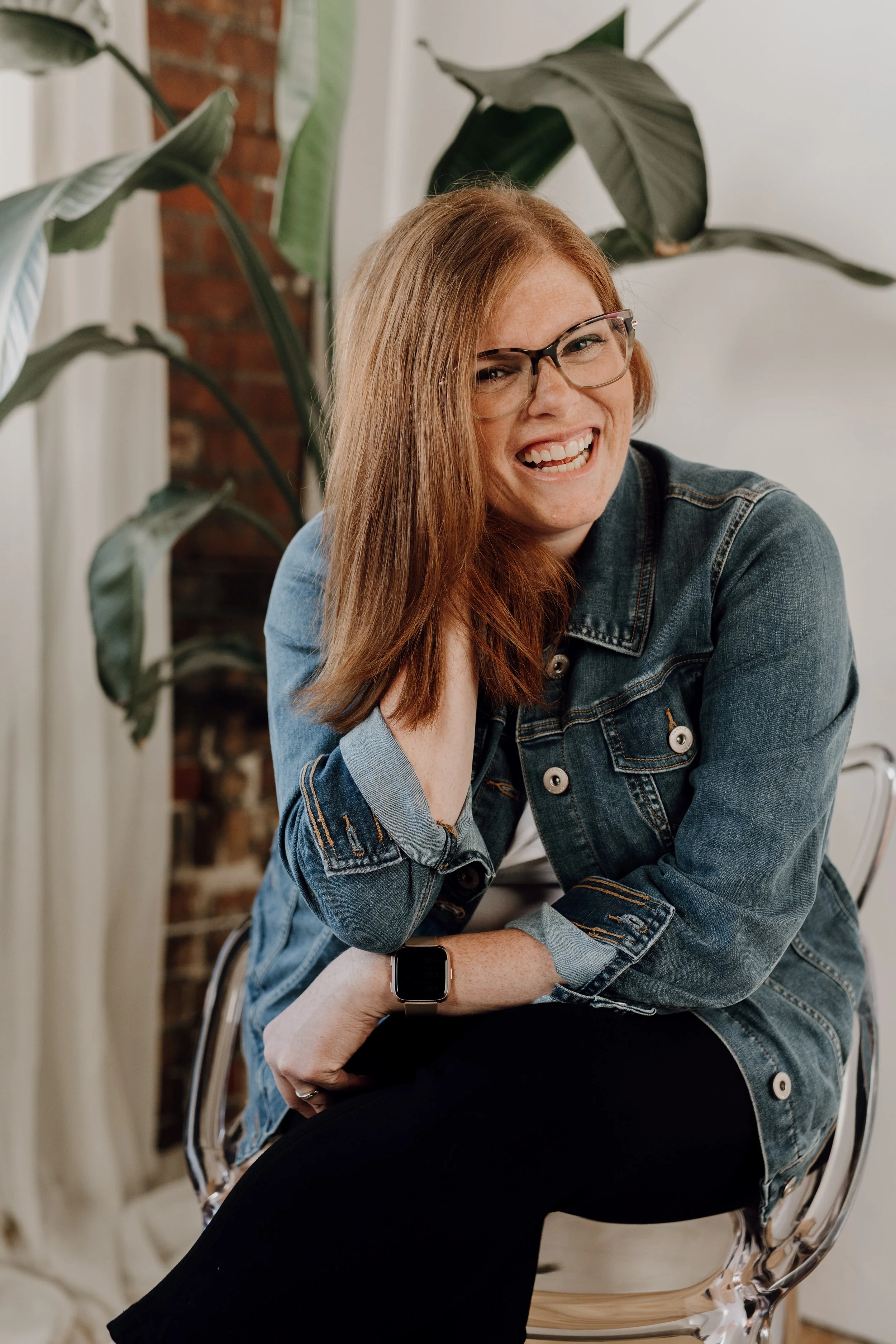 Allison, a glasses wearing redhead, is sitting in a clear chair laughing with a tall plant behind her. She's wearing a denim jacket and black pants.