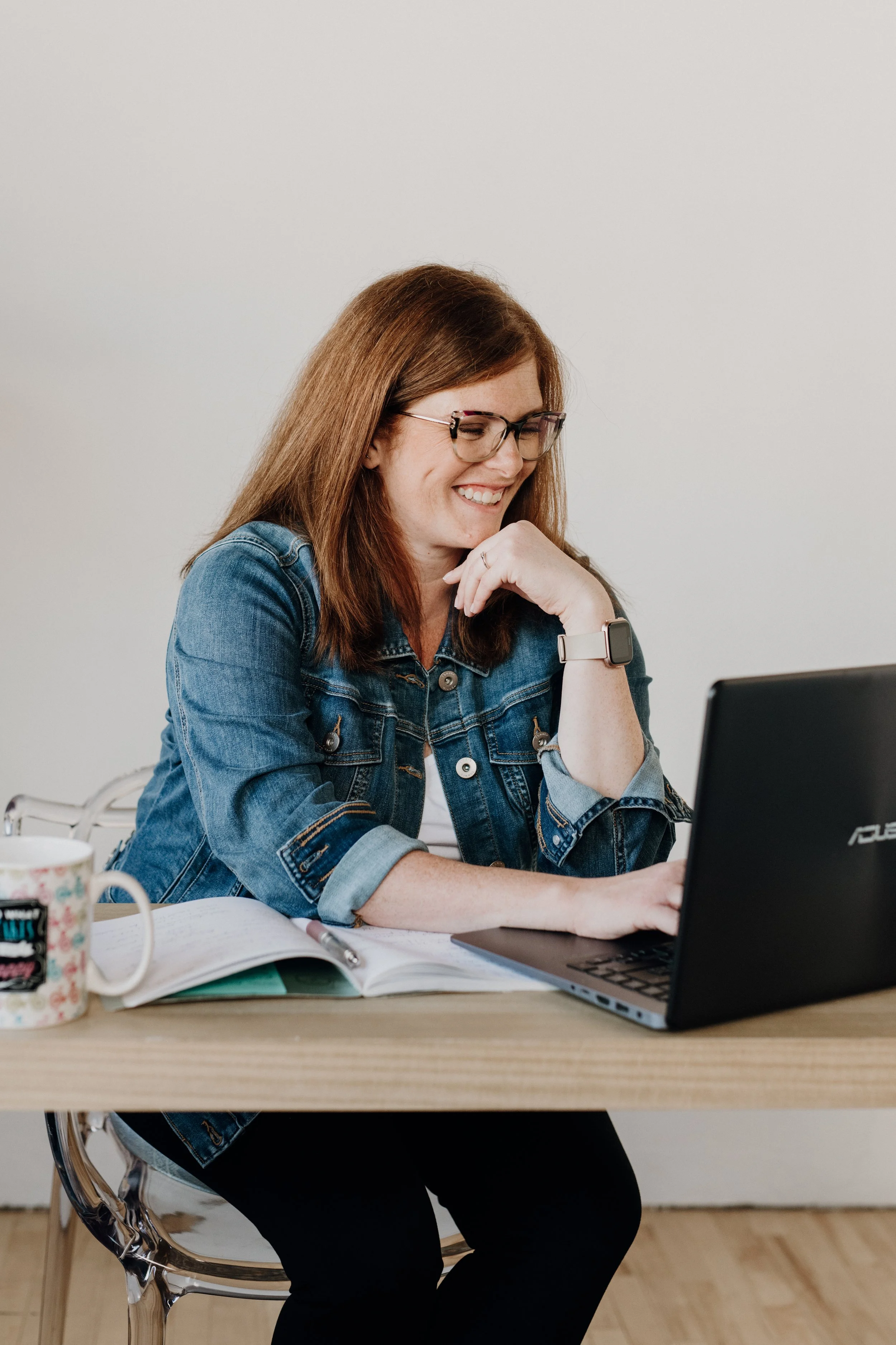 Allison, a glasses-wearing redhead wearing a denim jacket and looking at a computer while smiling.