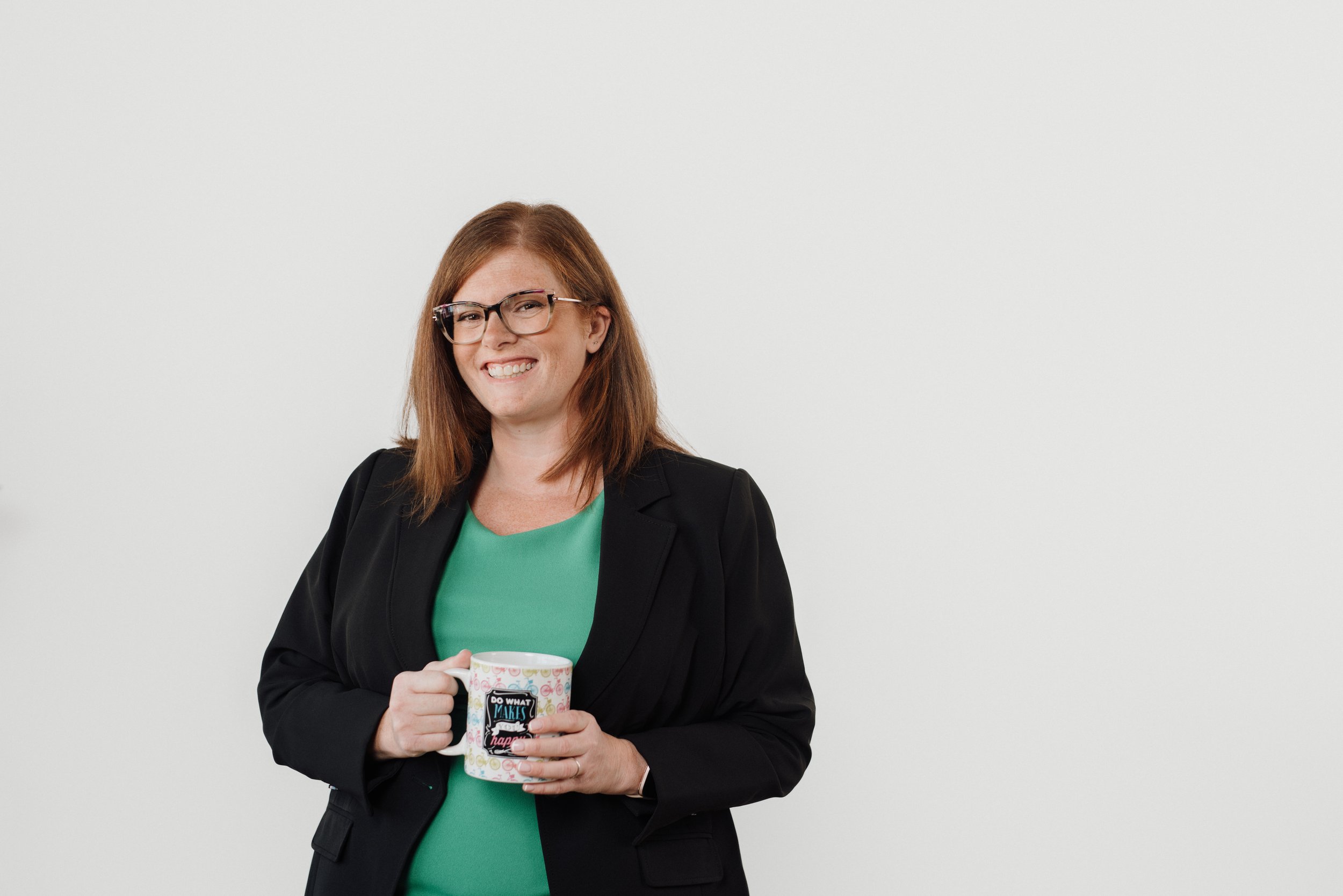 Allison, a glasses-wearing redhead in a green dress with a black blazer, smiling and holding a mug that says "do what makes you happy".