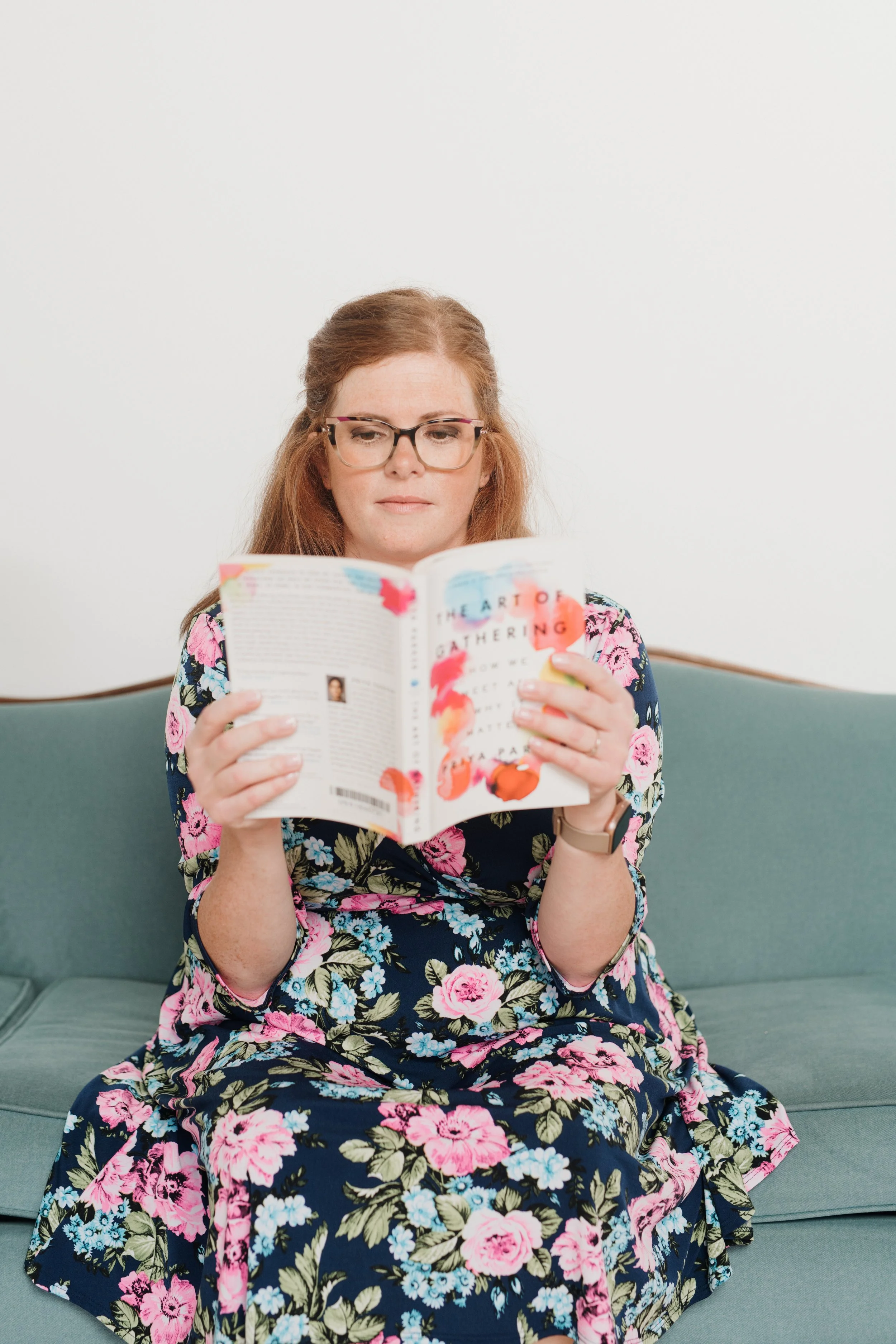 Allison, a glasses-wearing redhead sitting on a blueish couch, wearing a colourful flower dress, reading The Art of Gathering by Priya Parker