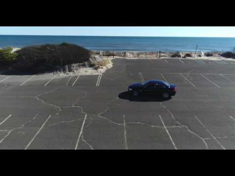 Aerial tracking of BMW through massive empty parking lot with ocean and beach in the background