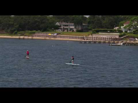 Low aerial tracking shot of two female paddleboarders, with beachhouses in the background.