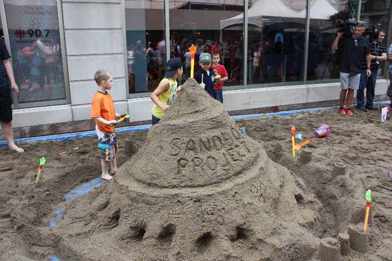 Some of the kids stand next to a sandcastle bigger than them