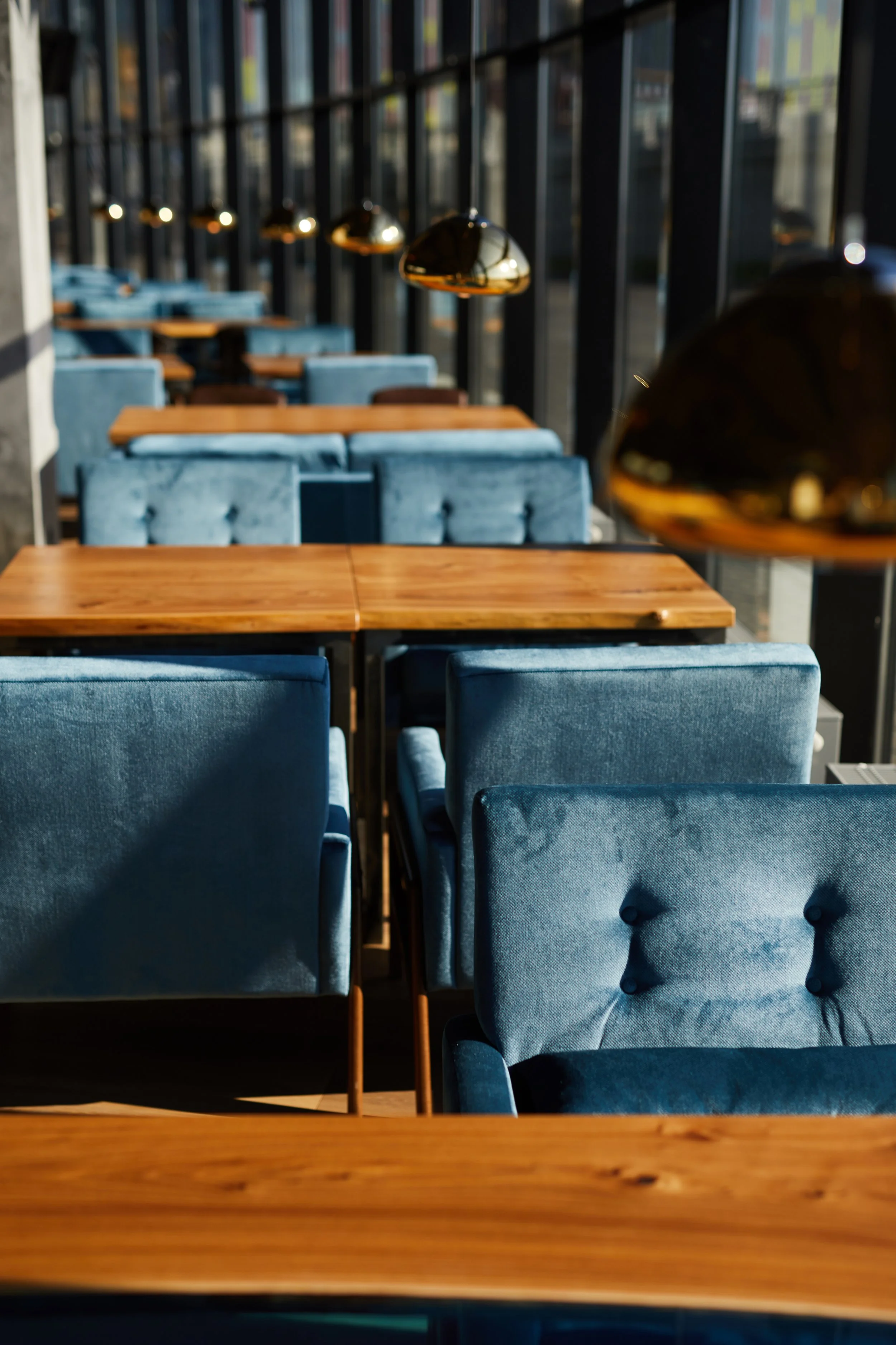 Contemporary commercial interior featuring rich blue upholstered seating, warm wood tables, and coordinated black and gold pendant lighting along floor-to-ceiling windows.