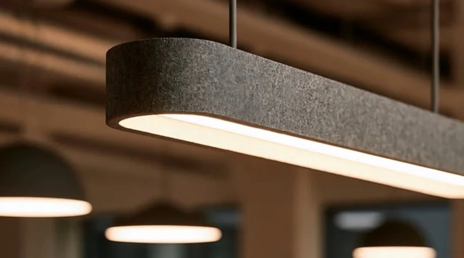 Close-up of a contemporary elongated pendant light with a textured grey exterior, softly illuminated underside, and minimalist suspended design.