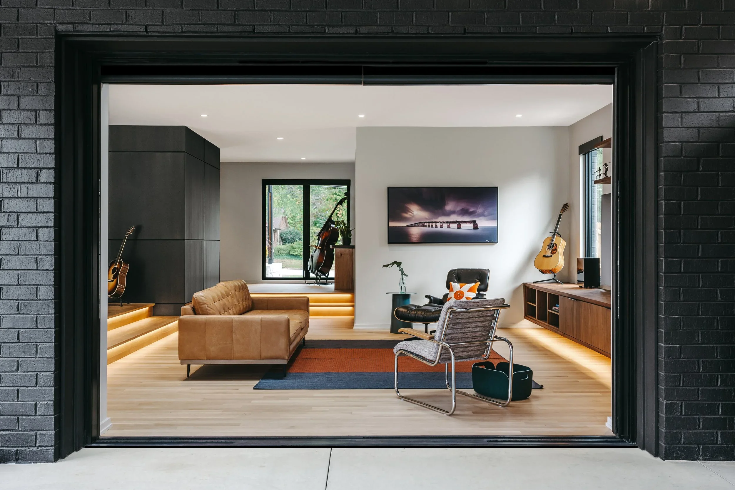 Modern living room with leather sofa, Eames lounge chair, and guitar, featuring a custom 20-foot floating black walnut media center with grain-matched cabinetry, integrated storage, and under lighting by Sunnyside Millwork