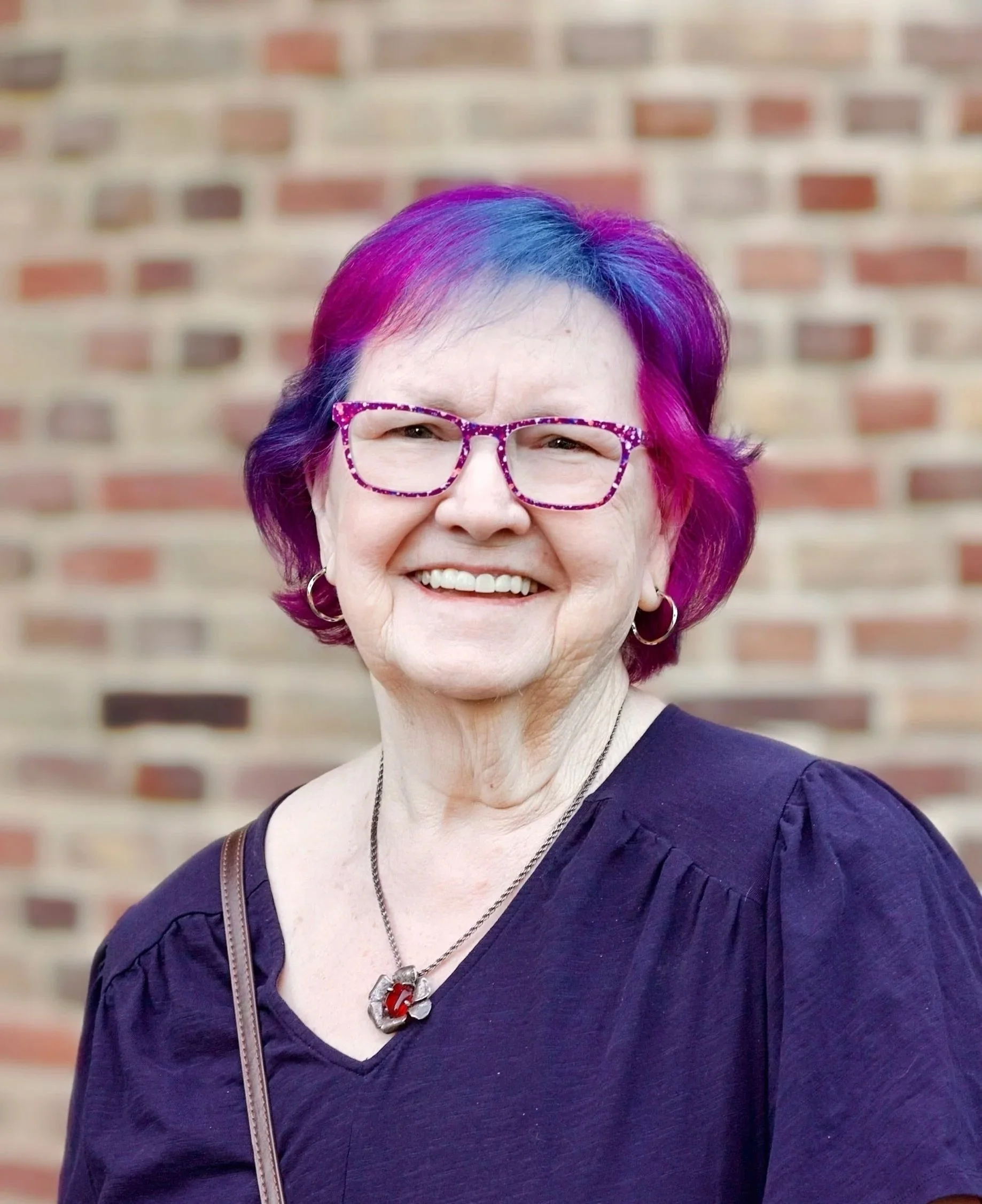 A smiling woman with short purple and pink hair, wearing glasses with a pink frame, hoop earrings, a necklace with a red pendant, a purple top, and a brown strap, standing in front of a brick wall.