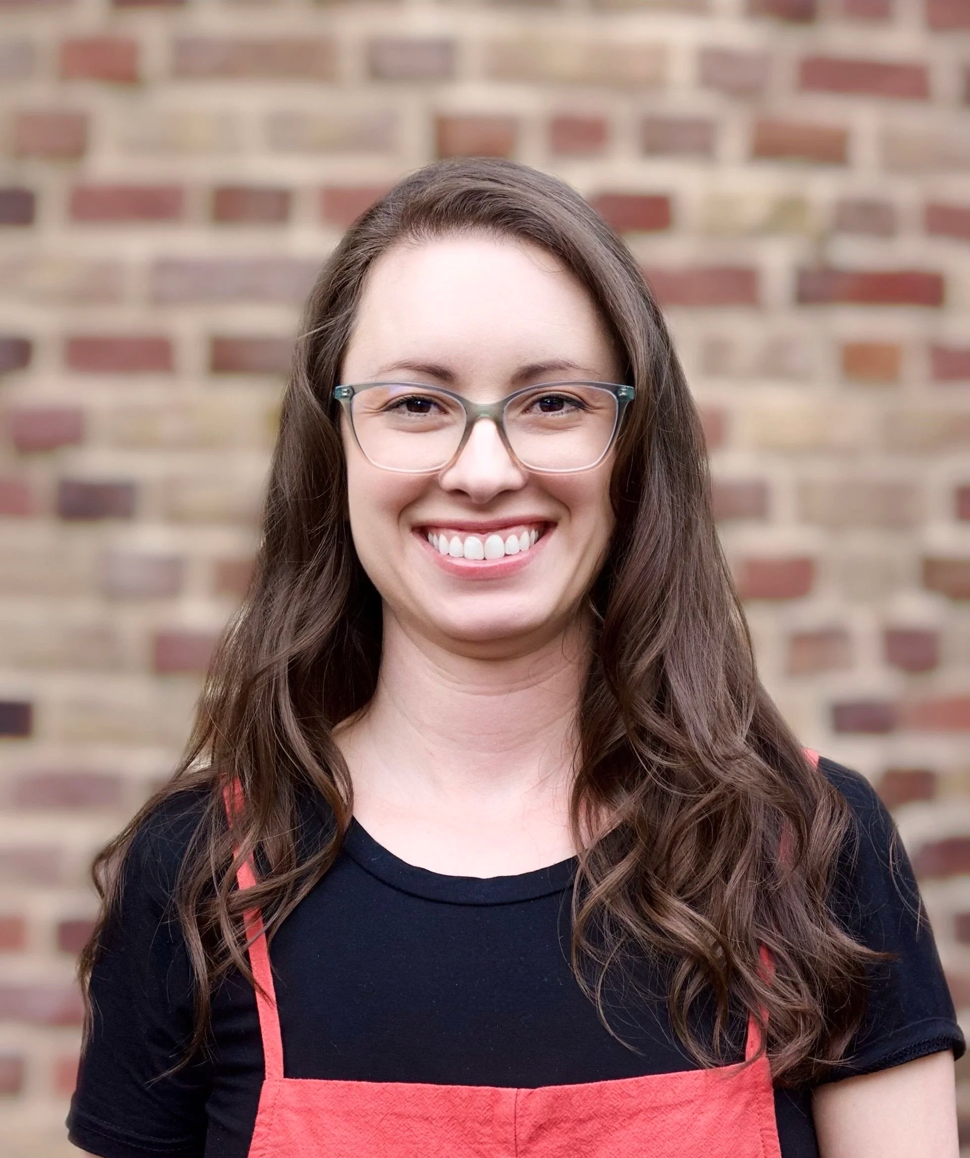 A woman with long curly brown hair, glasses, and a bright smile, wearing a black t-shirt and red overalls, standing in front of a brick wall.
