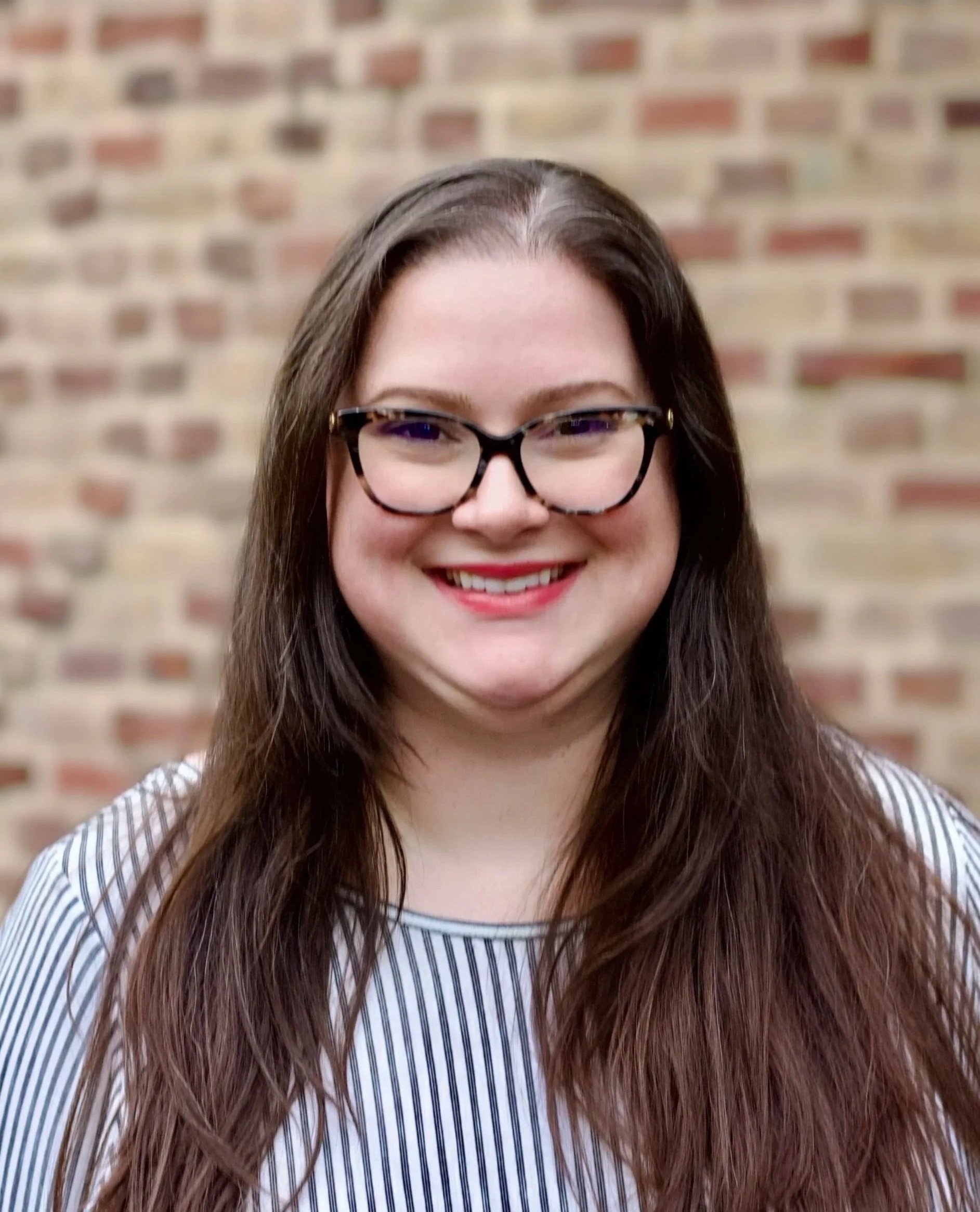 A smiling woman with long brown hair and glasses, standing against a brick wall background.