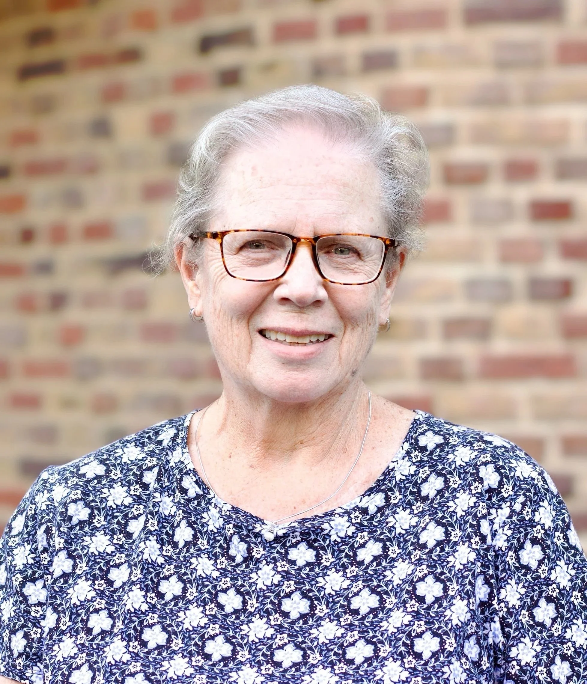 An older woman with gray hair and glasses smiling outdoors in front of a brick wall.
