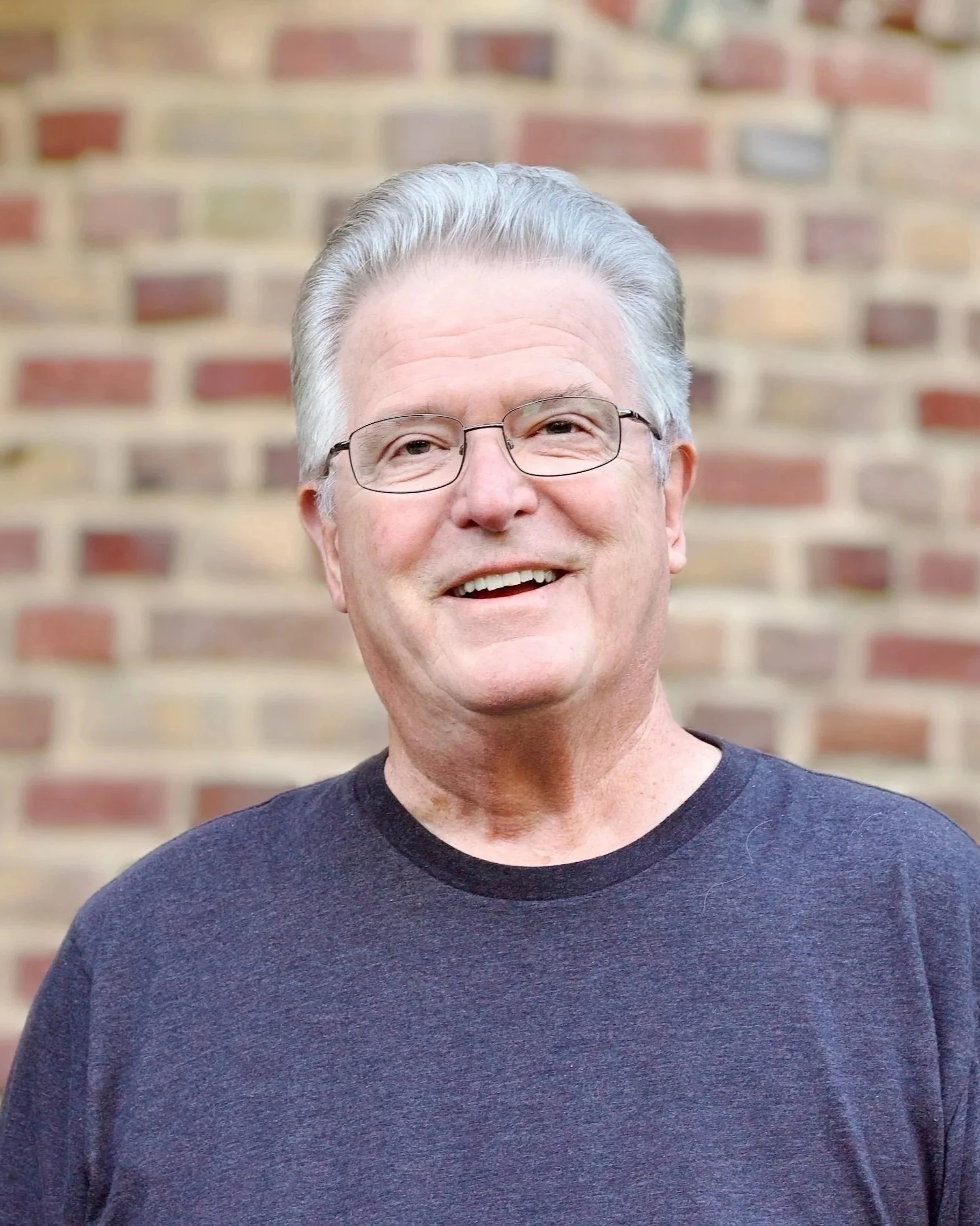 An older man with gray hair, glasses, and a friendly smile, wearing a dark T-shirt, standing in front of a brick wall.