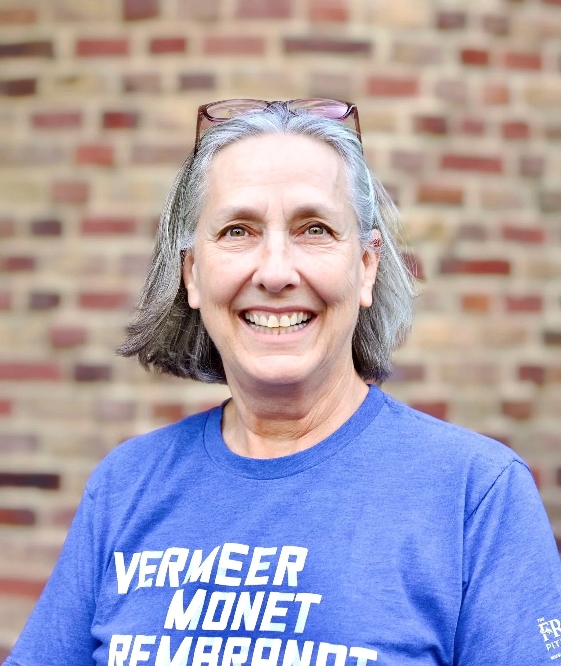 A smiling older woman with short gray hair and glasses on her head standing in front of a brick wall. She is wearing a blue T-shirt with white text that reads "Vermeer Monet Rembrandt" and other smaller text.