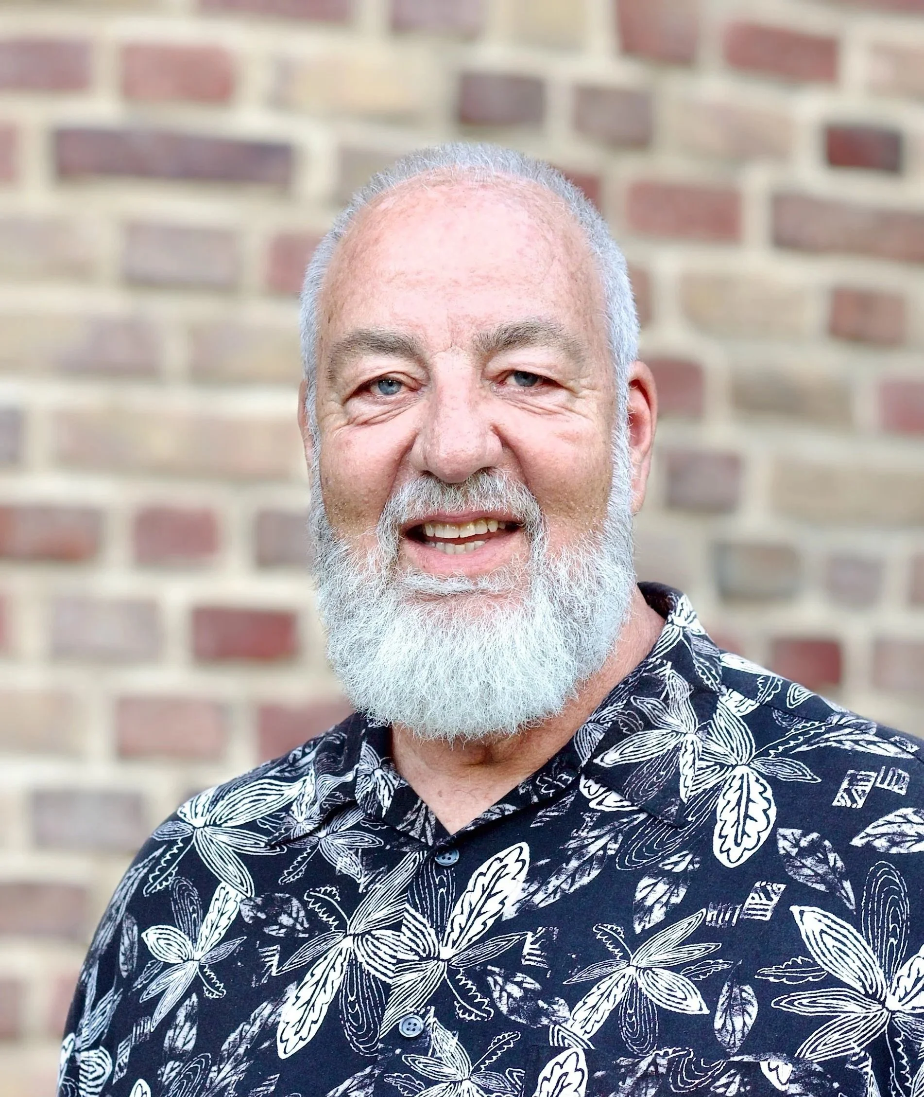 A close-up of a smiling older man with a white beard and gray hair, wearing a black shirt with white leaf and insect patterns, standing in front of a brick wall.
