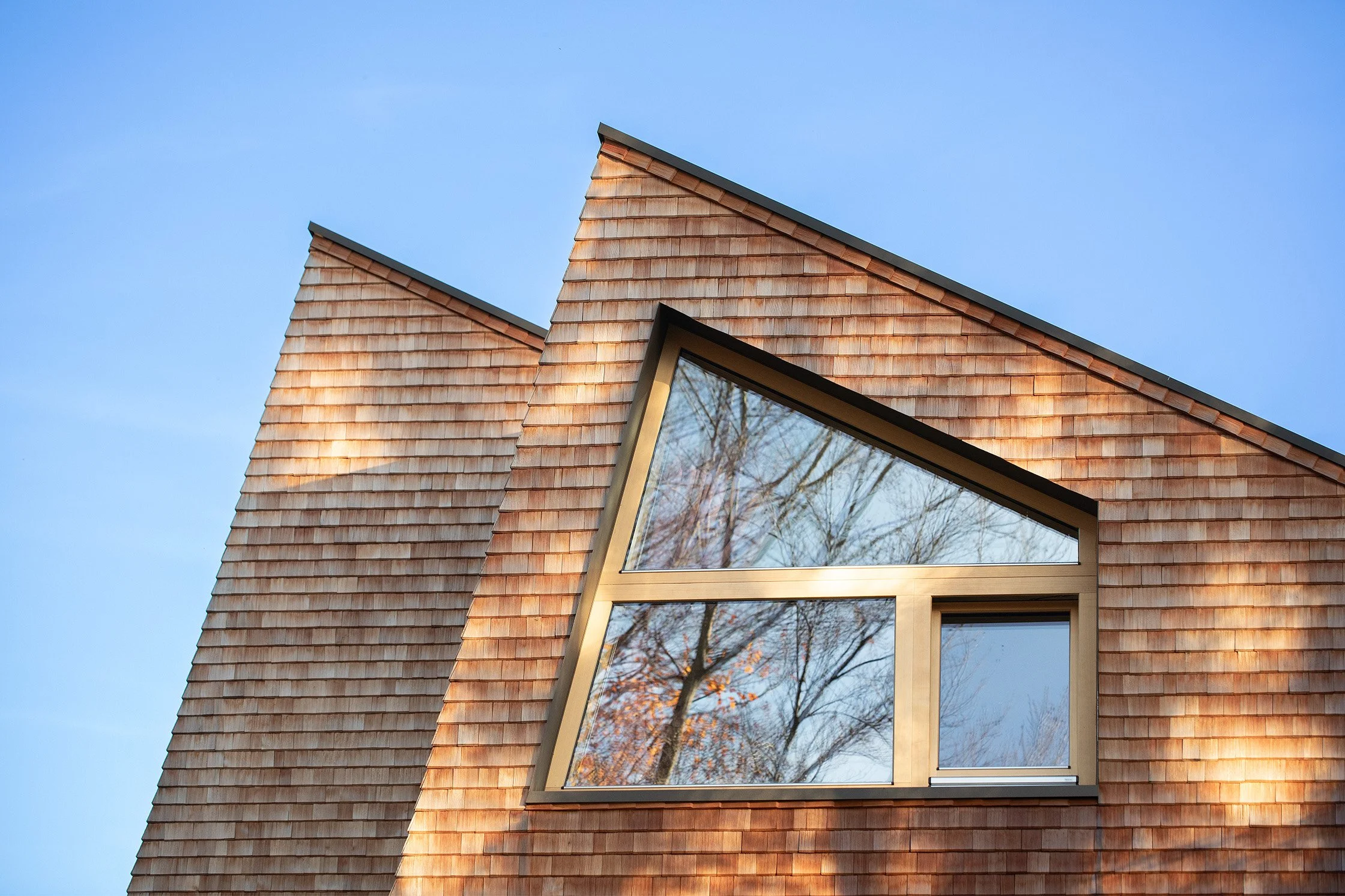 Detail der asymmetrischen Dachform von Haus W mit Lärchenschindeln und großem dreieckigem Fenster, das die besondere Geometrie des sanierten 1960er-Jahre Hauses hervorhebt.