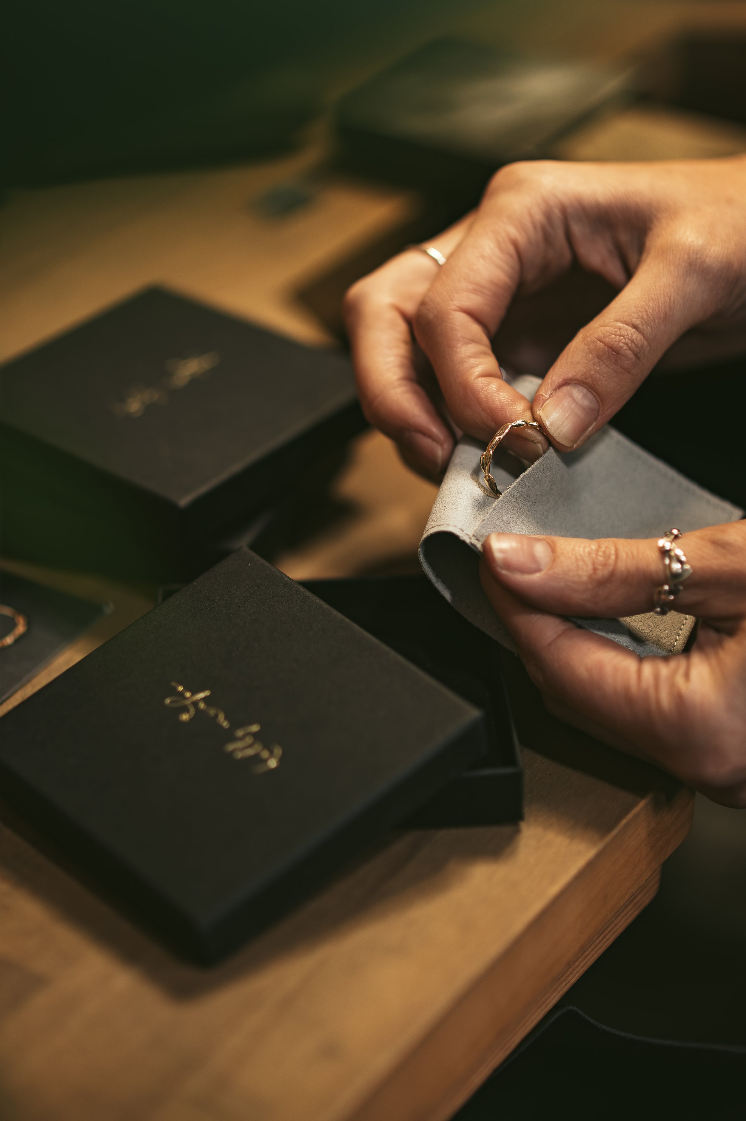 A close up of hands, carefully packaging an 18ct gold alternative wedding band into a pale grey, vegan suede pouch. Also shown are matte black, slim jewellery boxes, with Holly McAfee embossed in gold on top.