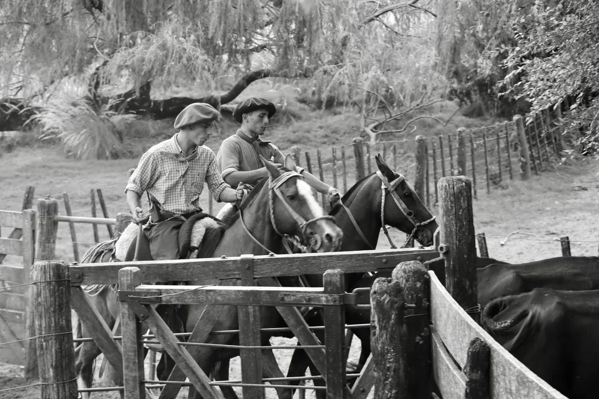 Two gauchos on horseback wearing traditional berets, leading horses through a wooden cattle gate at Estancia Los Potreros