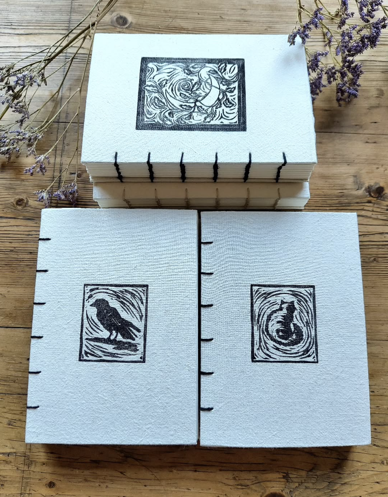 A stack of white books with black coptic binding sit on a wooden table with some dried flowers next to them. One cover featured a linoprint of the moon, one a linoprint of a crow and one a linoprint of a cat.