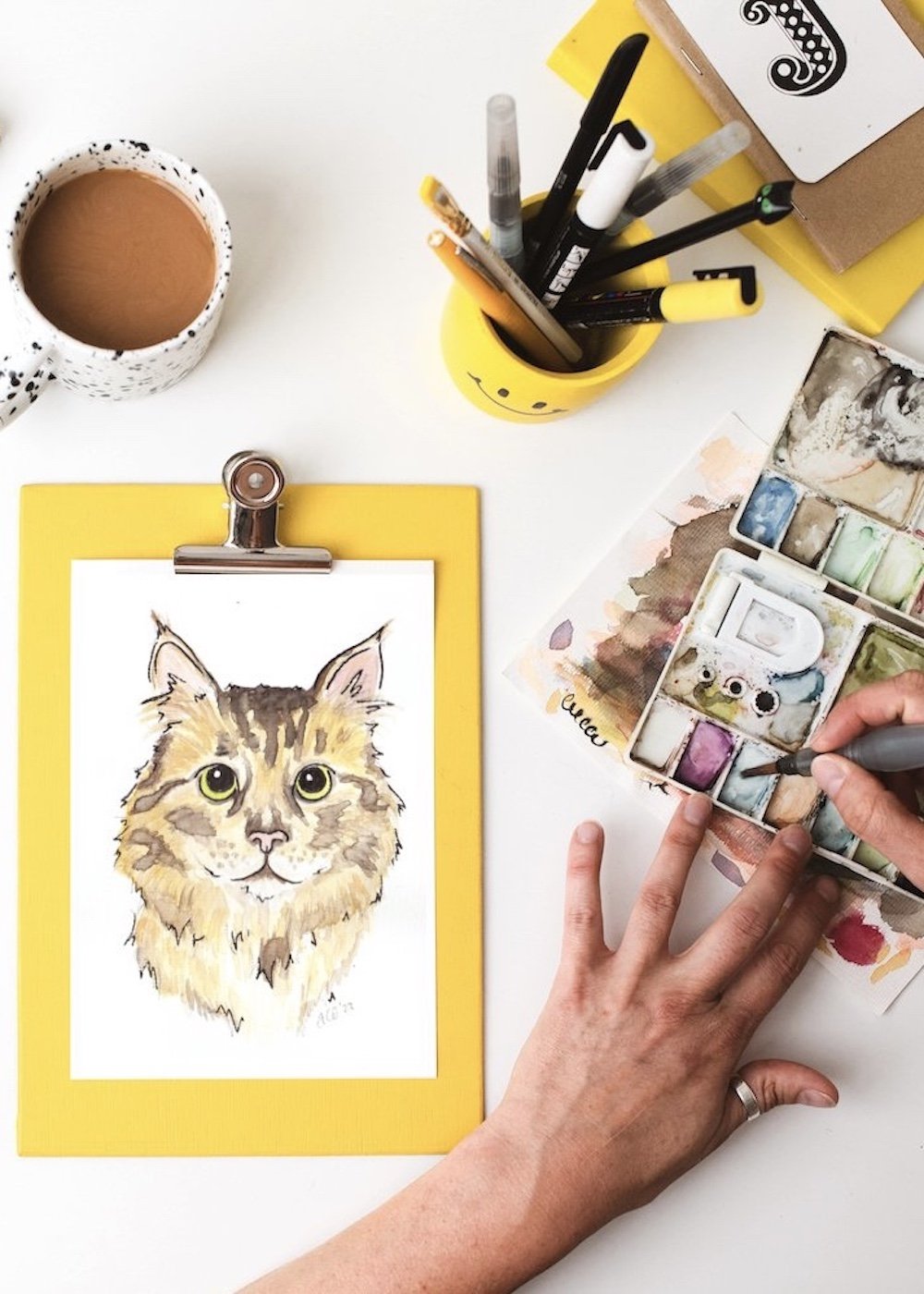 An overhead shot of a handpainted wateroclour portrait of a fluffy tabby cat. Jo's hands are in shot, reaching for her paints. Above is a mug of tea, a smiley yellow pencil pot full of pens and brushes and a stand of notebooks with a J on top.