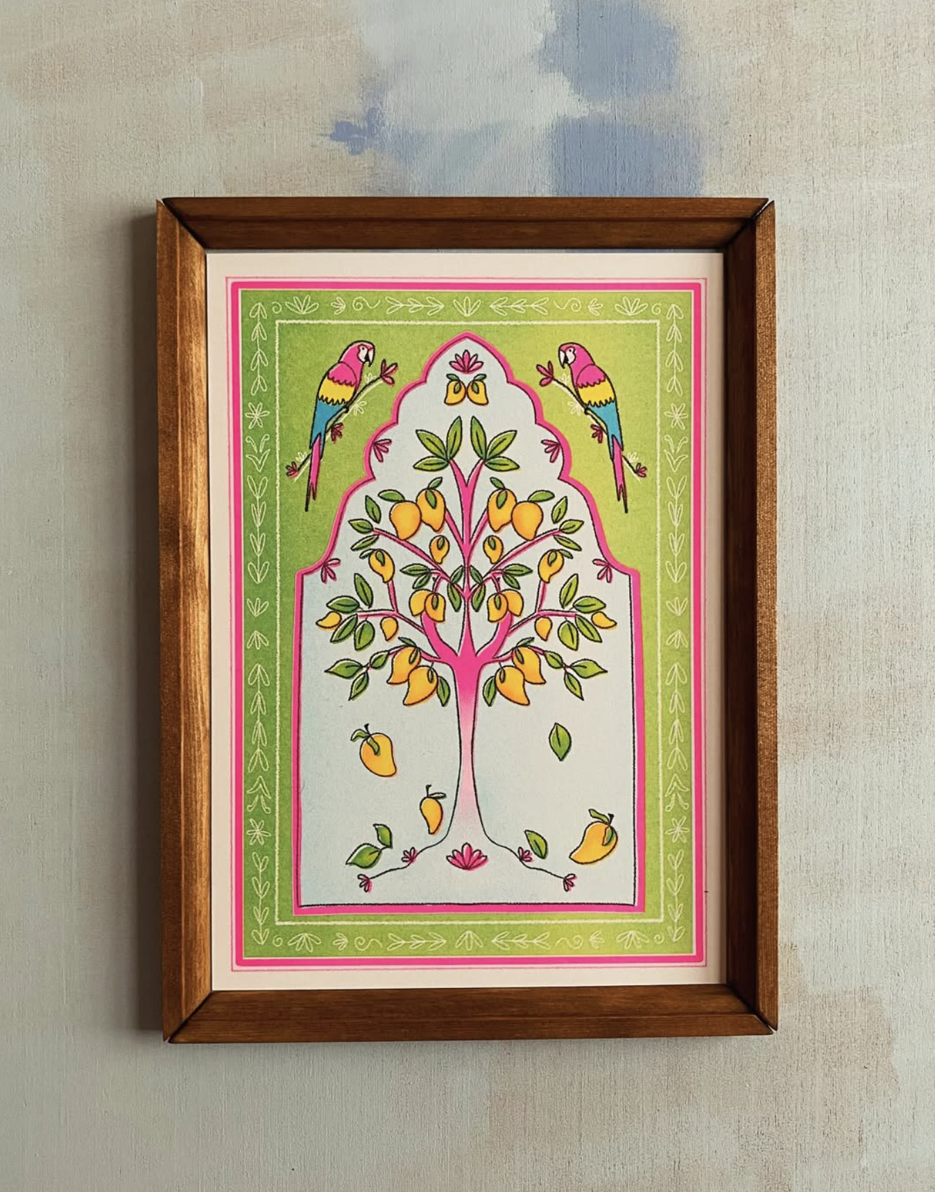 A framed print in of a mango tree in shades of green, yellow, blue and neon pink. A parrot sits either side of the tree with a green and pink patterned frame.
