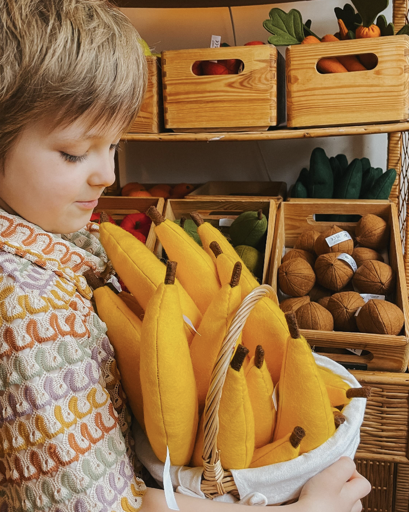 A young boy with blonde hair and a patterned shirt holds a basket of felt bananas in front of wooden crates filled with other felt fruit and veg - pears, coconuts and carrots can be seen.