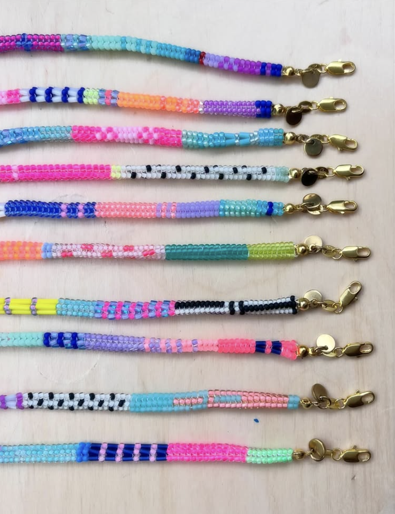 The ends of ten necklaces lined up - each is made with small brightly coloured beads and features a unique pattern. Each has a gold clasp at the end.