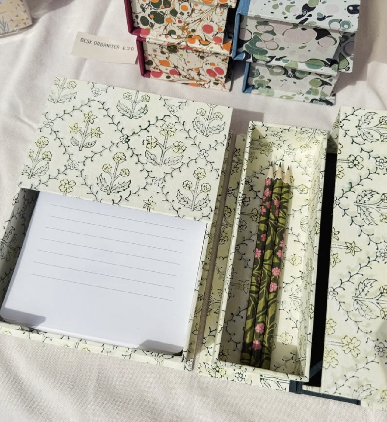 A cream and green patterned box lays open, showing lined notepaper in one side and three patterned pencils in the other. In the background, other handmade boxes made with marbled paper can be seen.