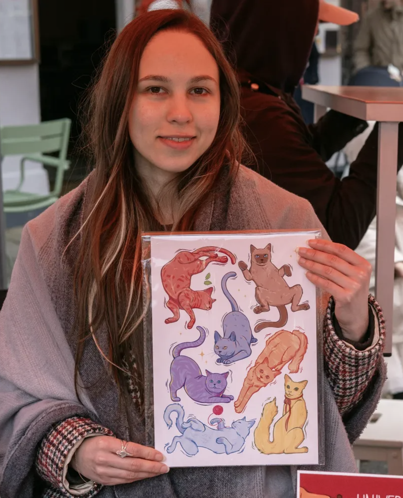 A white woman with brown hair and blonde highlights holds up an illustrated print of several cats in different poses.