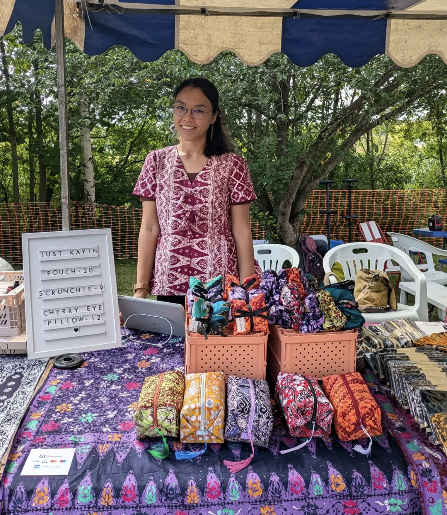 A Malaysian woman with tied back dark hair and glasses, wearing a maroon batik dress stands behind a market stall featuring patterned batik pouches, scrunchies and eye pillows.