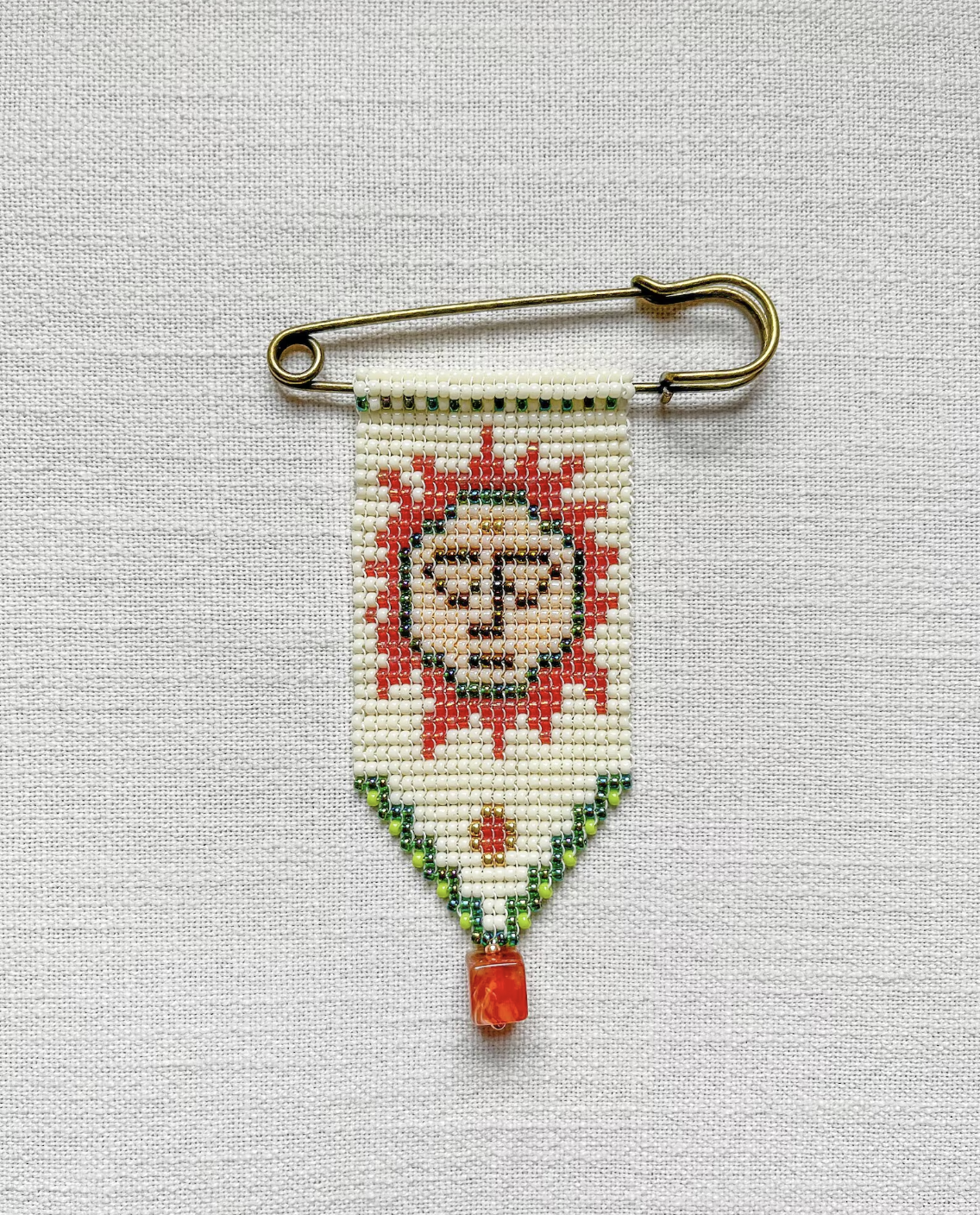 A beaded brooch featuring a large safety pin, on which hangs an intricate beaded pennant. The beaded design featured an orange sun with a face on a cream background, green patterns and an orange charm hanging off the bottom.