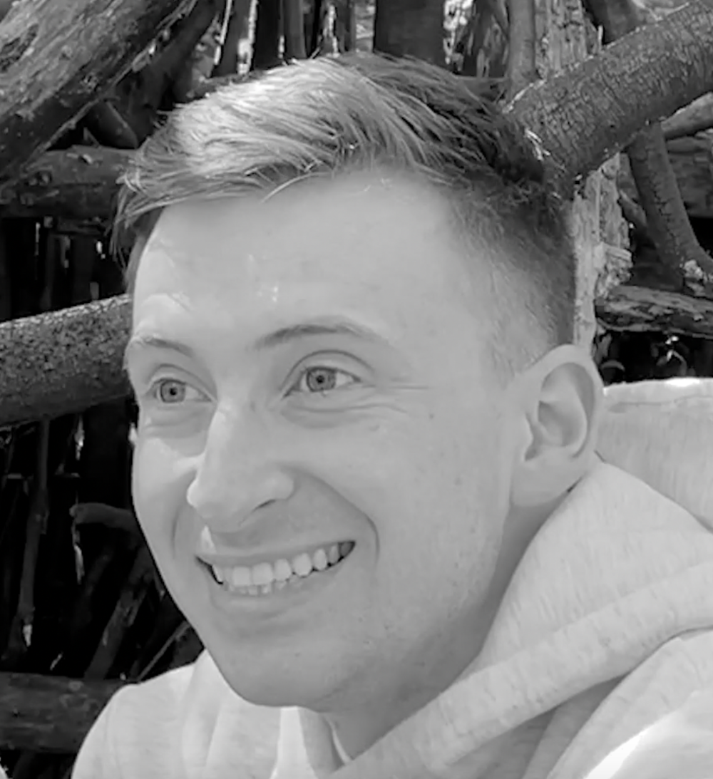 A black and white headshot of Sam Dunstan, a white person with short hair, wearing a pale coloured hoodie. Sam is smiling and there are tree branches in the background.