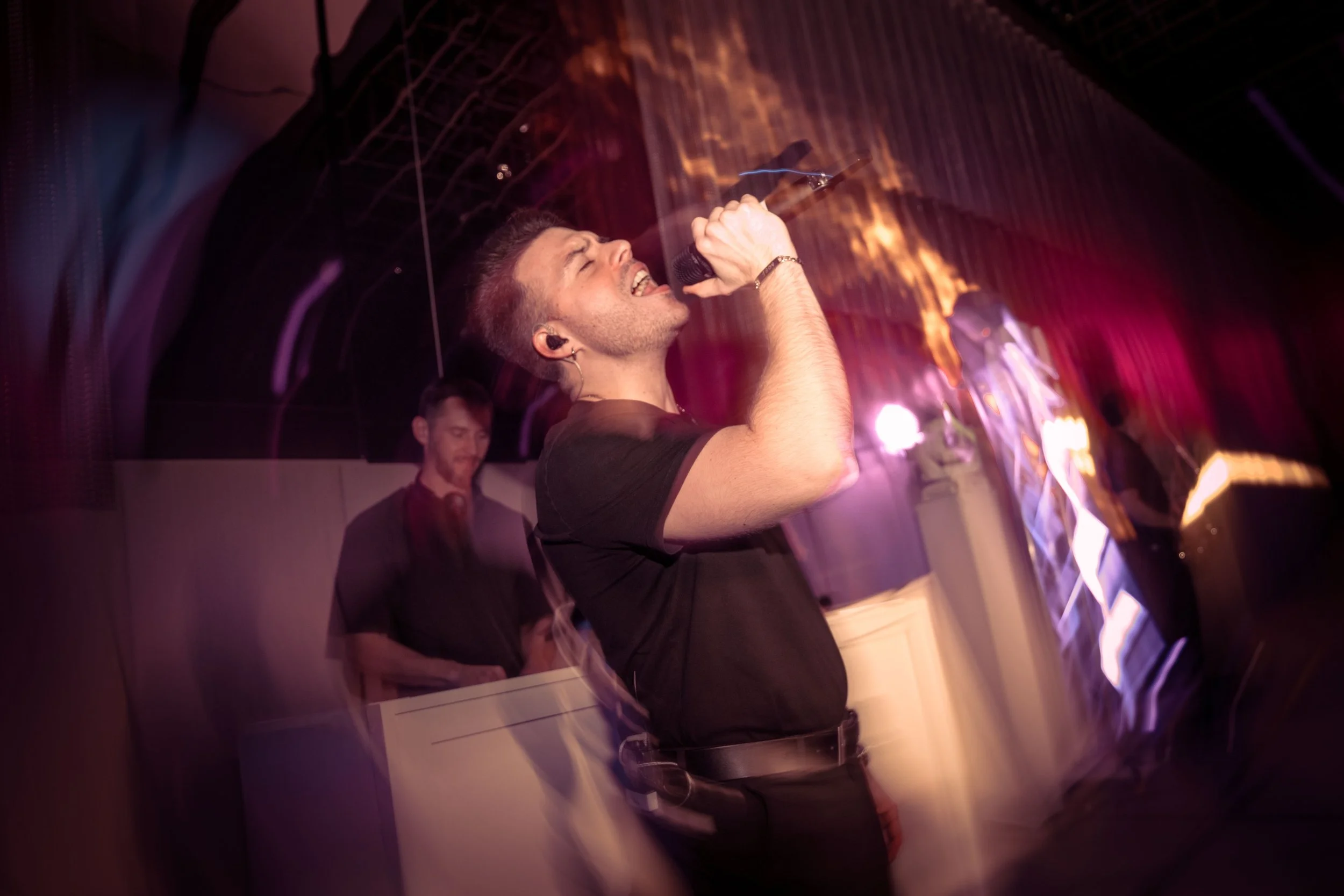 A man passionately singing into a microphone at a karaoke or live music event, with a DJ or music technician in the background under colorful stage lights.