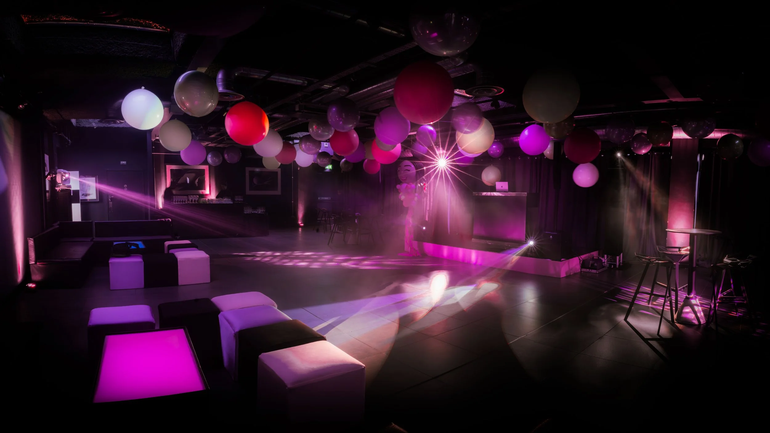 Empty nightclub with purple and pink lighting, decorated with hanging balloons and illuminated furniture, with a dance floor and a DJ booth in the background.