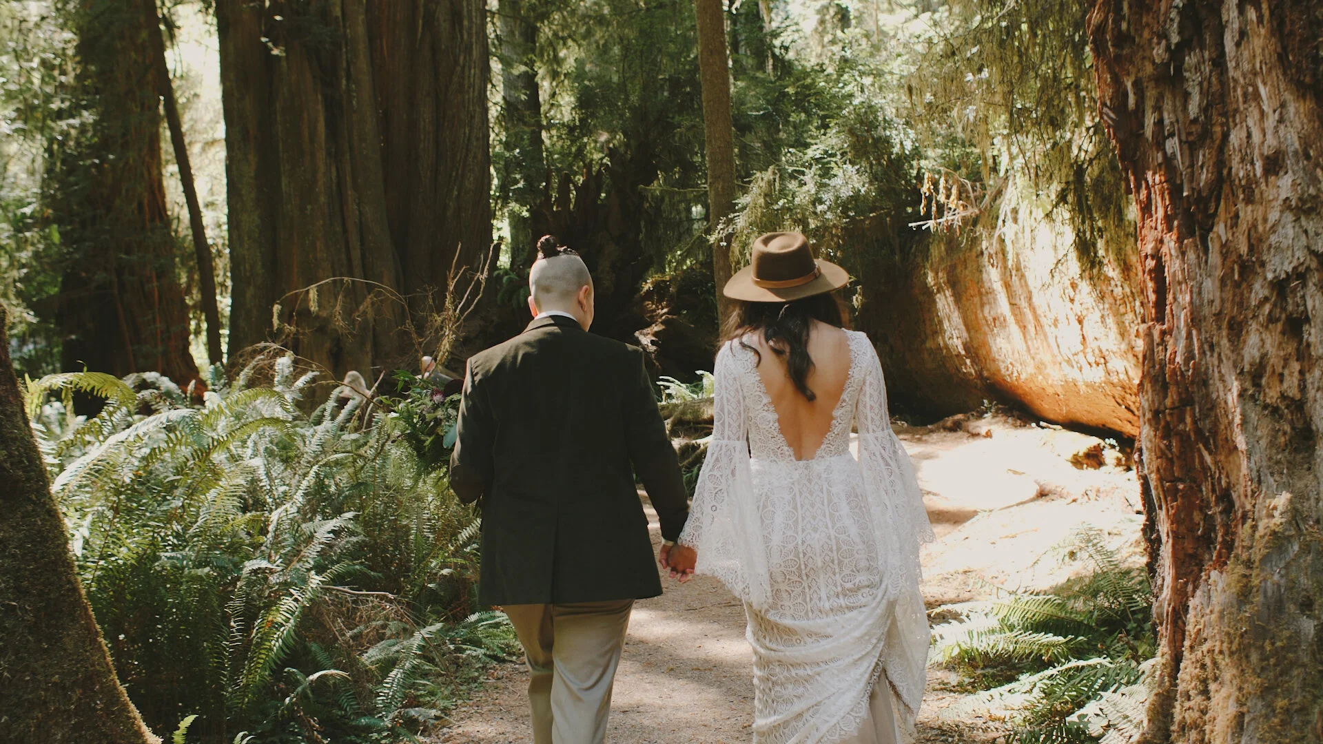 Couple walking through the redwoods after their wedding under the trees. They are holding hands and facing away from the camera.