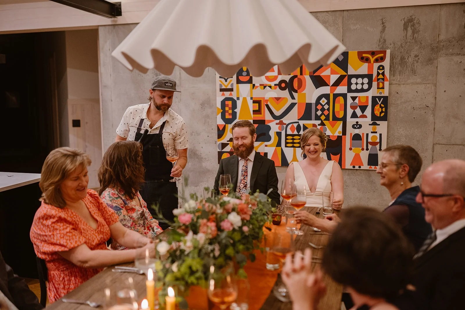 A bride and groom laugh surrounded by family at their modest wedding dinner party
