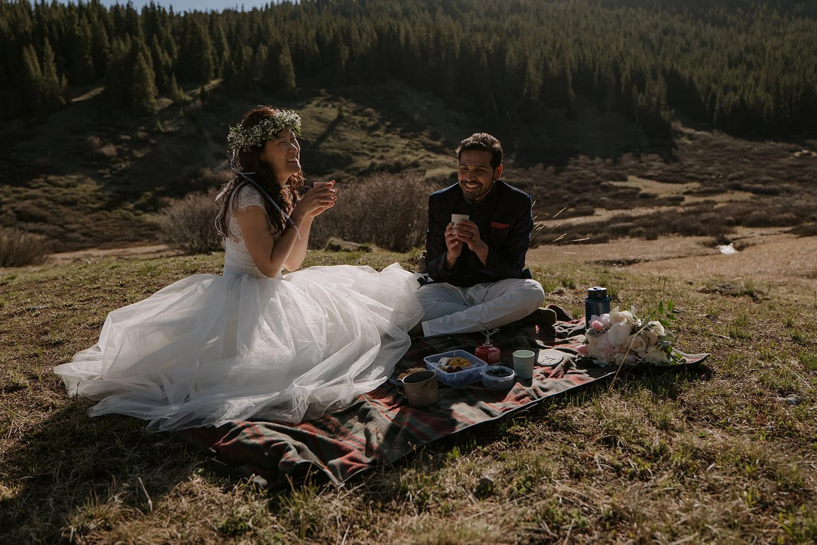 A bride and groom enjoy a rustic picnic in a sunny clearing