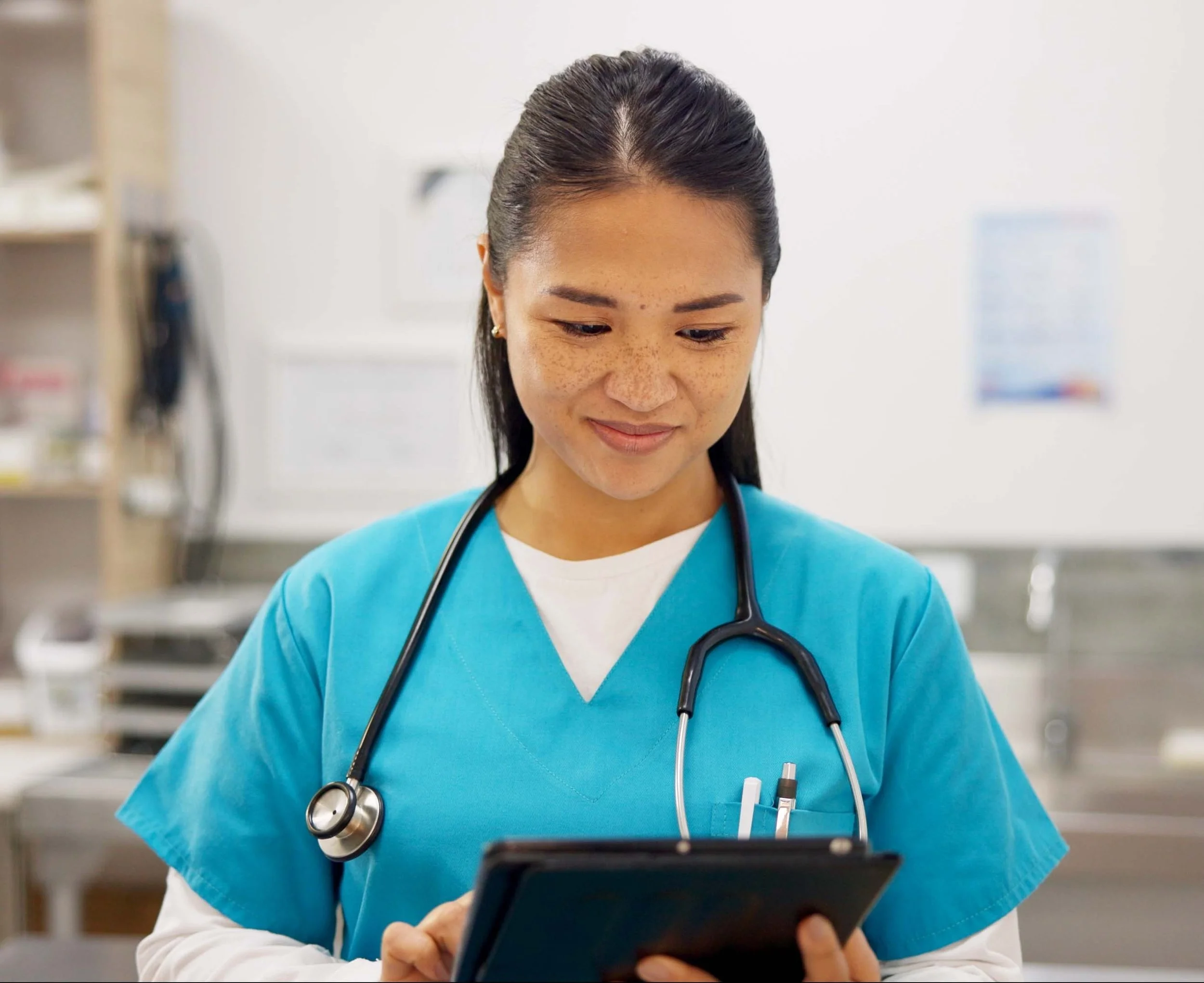 A smiling woman veterinarian with dark hair pulled back, wearing bright teal scrubs and a black stethoscope, looking down at a tablet in her hands while standing inside a clinic.