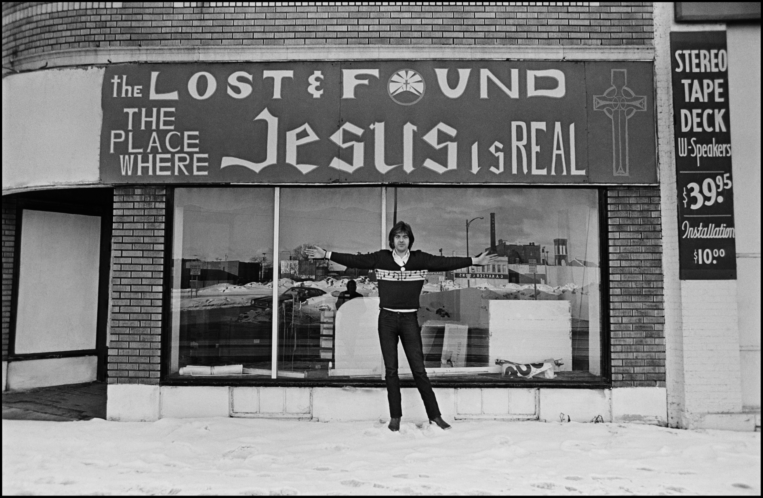 Nick Lowe - Buffalo 1978For any British band touring America in the 70's the first thing you did after checking in to the Hotel/Motel was to hit up the Thrift Shops and Pawn Stores. Back then there were still gems to be had at both, and musicians rigorously checked them looking for guitars and other instruments, on this particular day Charles Shaar Murray found a Telecaster in great condition, in fact he lent it to Nick so that he could play Heart of the City in the encore at Elvis's show that night at the University. Thrift Shops, also known as Sally Ann's (Salvation Army) were great places to find cool stage clothing, and also winter coats, much needed as this trip occurred during the great Blizzard of '78. These were the days before Johnson and Johnson started being the place for Rock Star Threads, us chaps could not get enough cowboy shirts, leather jackets, cashmere coats and a whole lot more. The Thrift shop in this photo is in Buffalo, and the temperature was 15 below, but Nick Lowe is a hardy chap and never even bothered to wear a jacket over his legendary musical note sweater.