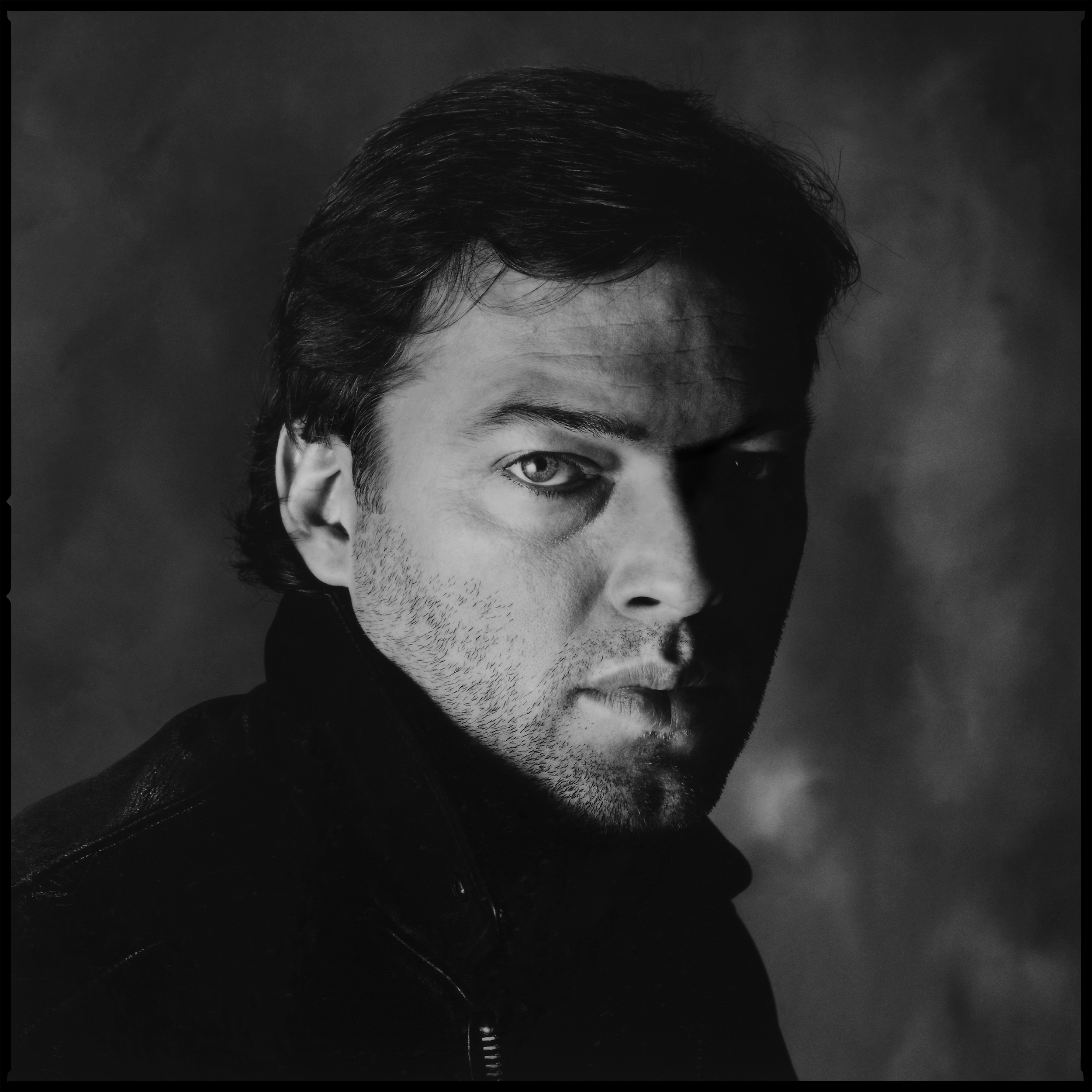 David Gilmour - London 1983 When I received a call to see if I was interested in photographing David Gilmour I could not reply in the affirmative quickly enough. Not only that but it gave me the chance to work with Storm Thorgerson of Hipgnosis fame. Storm was the one who inspired me to become a photographer, the sleeve for Atom Heart Mother featured a cow, no band photos at all, how cool was that. I fantasized about being the photographer, what a great way to earn a living, I was only 15 but my mind was made up, this is what I wanted to do. The chance to work with Storm and David 12 years later made that dream come true. The word came from on high, "Tell Chalkie to get a Water Cooler" but we don't have them in the UK so I told him no. He refused to budge, we got one from America, I asked why? He said I have got this Photo, trust me. We were booked into a white background studio, I could not fathom this water cooler bit, but, whatever he wants I'll do it. The Water Cooler arrives, we fill it with water, we wait three hours for Storm to show, I ask to see this picture, he hands it to me. It was James Dean, outside a store, leaning on a Water Cooler. He was radiating handsomeness. My heart dropped, David's not used to this studio photo stuff, no way can I turn him into James Dean instantly. And Storm wanted to start with this. But I was touched by the fact that he thought I could turn his friend into James Dean, that David could become James Dean. David is David, he's very special, but I could not possibly do that without teaching him how to act in front of the camera, not just be himself, and teaching him that would take a few hours. Storm starts ordering everyone around, barking orders at the crew, they get nervous moving the cooler and knock it over, the glass shatters, the studio gets flooded. I'm relieved and smile at David, he smiles back and opens a guitar case, out comes the Red Strat and he sits in this cheap plastic chair and entertains us for 45 minutes while they mop up the studio. Starr takes an SX70 Polaroid of him doing this, in my friend Scott Crolla's groovy shirt, then we realize he needs stubble, he's so clean-shaven that he lacks his look, he has none of that slight grubbiness he needs, so I tell Storm and we cancel the whole thing. Just one SX70 had been taken, thousands of pounds had been spent. We regroup in the Canteen at Abbey Road, figure out the growth time, then it came to the crunch, I had to persuade Storm not to come to the reshoot, he needed to trust us to do our work in private, our little portrait studio was in the garage of a Mews House, it was tiny. David came, Starr did his make up this time and nobody else but Storm's trusty Ambassador, Andrew Ellis was present, It features the difficult photographic technique of lighting from below, needed to cast a slight shadow up onto his cheek and to generally add a bit of drama, he's such an incredibly nice and handsome person that I felt we needed to make him a little tougher looking. We gave him my battered old leather jacket, put him on a chair and went in close on his face, he happened to move his arm quickly and his hand crossed his face and looked like he was thumbing a lift, Starr managed to catch this frame and ultimately it was used as the cover. She liked people smiling, I liked them moody, so the portrait you see here went on the inner bag and the happy one on the cover, after all David is a pretty happy kind of chap, so it fitted in well. It was also pretty cool to have his Electric Blue Porsche parked outside our crummy garage for the afternoon. One of the things that comes as a bonus when photographing guitarists is that you can ask them to bring a couple of instruments with them, it does two things, it gives them something to hold and covet in front of the camera, it also gives them something to do when backgrounds and lights are being changed. And they do what they do best, they play their guitar. It's a very privileged thing to hear them noodling about whilst they are waiting, and, you get to see some pretty amazing guitars too. Also worth mentioning is that when he reformed Pink Floyd seven years later with my dear friend Guy Pratt replacing Roger Waters on Bass, they Toured the US after a month of rehearsals in an empty Aircraft Hanger at Toronto Airport. Sitting in a pair of Directors chairs Storm and I watched the final night of rehearsals with the full Stage Show including Lasers, the band played a two hour set to an audience of less than ten people. To say it was a mind blowing experience would be understating things, the best bit was during the encore when they played Comfortably Numb, with it being the last night they took a chance and opened the Hanger doors just as David got to the guitar solo, the lasers projected out into the night sky creating an incredible light show, then, just as the song was finishing a Jumbo Jet went by and the Floyd's stage set was dwarfed by the size of this massive plane. The reason for the tiny audience was that in order to get to the gig you had to be driven across the runways at Toronto's Pearson Airport in a special vehicle, it had two huge flags sticking up from the roof in order to make it visible to the Pilots of all the Jets that were taking off and landing, obviously that's a highly restricted access route so it meant very few people other than the Band and Crew could get there...