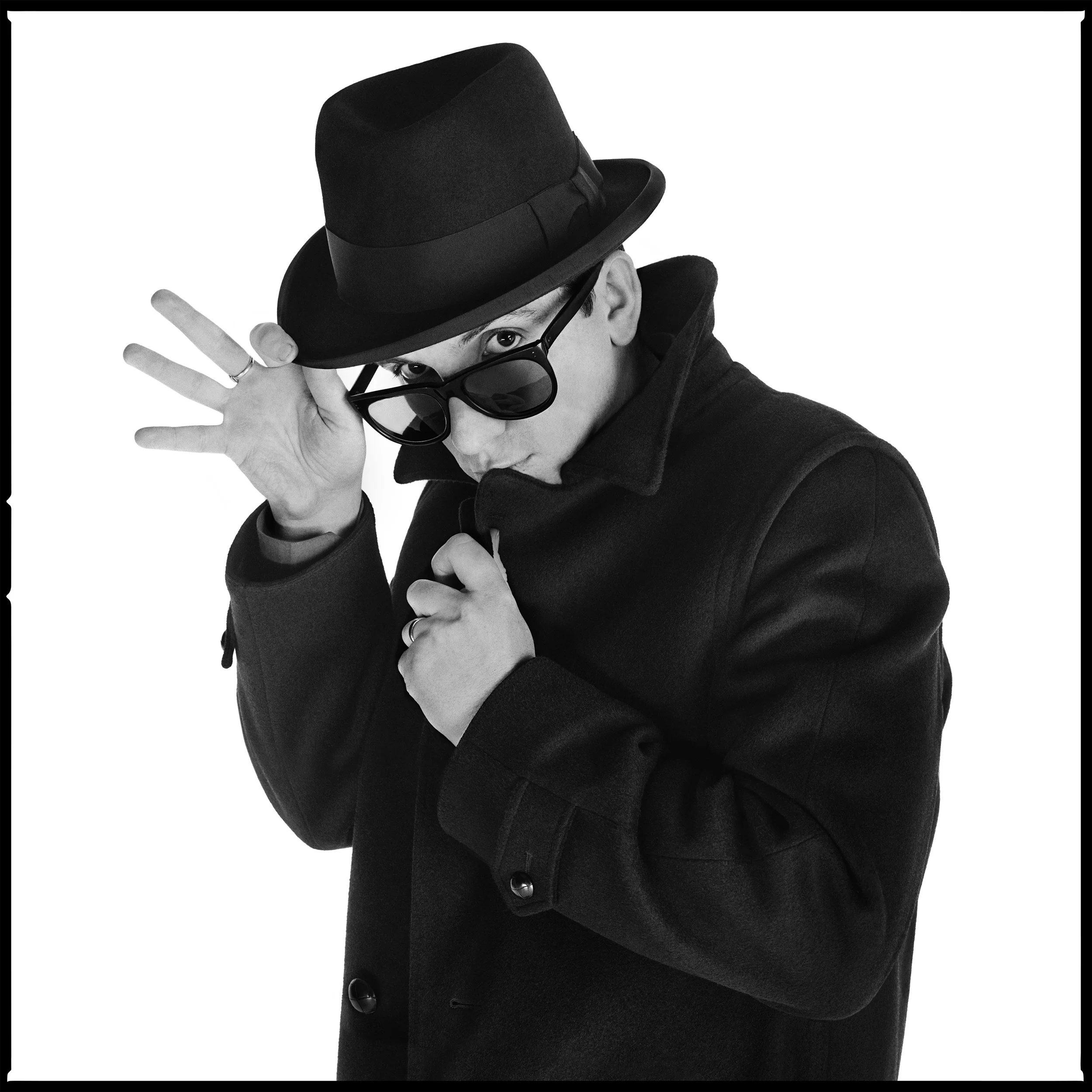 Elvis Costello - The Trust Sessions 1981 Elvis Costello was the first person I ever photographed in a Studio, it was March 1978. We did it at the same time as the two consecutive NME Cover stories, one with Nick Lowe, the other of him sitting on a bench. I never showed them to the paper, mainly because they were in color, but also because Studio Photography was not allowed yet, documentary, live, or while they were being interviewed was the norm back then. I set about changing that, but I had to wait until 1980 and the launch of the Face before anyone took me seriously, Pop Stars on White was simply not considered interesting, it was considered to be boring, the stuff for PR photos. But in 1979 Jake Riviera asked me to shoot EC in the Studio for Armed Forces, he had a vision of Elvis smoking, I had a vision of him standing with his guitar, his feet awkwardly bent, but in the end a painting of a Herd of Charging Elephants was used, Jake's smoking photo was put on ice, and the full length shot was printed 70 feet high above the Dominion in Tottenham Court Road. Trust was our next venture into the Studio, this time it was somber, dark hat, coat and suit, a cigarette in hand, it got proofed, but in the end it lost out to a frame from Barney Bubbles Video for New Lace Sleeves, a color shot of EC looking over his glasses, it became somewhat Iconic. The pose was something we came up with in the Studio, weeks before the video. That image had a Frank Sinatra vibe to it, his hand making the perfect wave, I loved it but it was not part of the brief, so it appears here for the first time. Next up was something that was the ultimate photographic honor, the chance to use the giant 20x24 Polaroid camera, that produced an instant print two feet high, there were only five of these cameras in the world. I called EC and asked him if he would sit for me, he eagerly agreed, the camera was incredibly slow to use, and you only got ten sheets of film, I shot nine frames, with only three decent photos, one of which appeared on the cover of the Face, the last frame however was something completely different. He looked away from the lens, I asked him to look down, wearing his black beret Jake joked that it was his Laurence Olivier pose, in retrospect that was quite a compliment. We shot more pictures to go with the Face story, but it was 1982 and from his bag he produced a blue sweater, and matching blue glasses. I documented it, but kept it hidden, if Jake had seen that I would be in trouble, no way would he want EC to be photographed in that outfit. I thought it was cute, but it remained under wraps for 32 years. In 1986 we worked again, around the time of King of America, as well as some Django Reinhardt looking ones, EC suddenly appeared in a bright yellow coat, and black watch tartan shoes, I made him jump up and down, jumping for joy was the Fleet Street name for it. In 1991 he teamed up with the Brodsky Quartet and I got to shoot him with Classical Musicians, they were about to go on tour, I warned the musicians of the perils to come. You are going to get screamed at, way more than the polite applause you get at Classical a concerts, it will change you forever, you will never be the same again, I was right. Although I had given up photographing people by the mid nineties, I did agree in 1996 to do one last session in New York, to promote North, his latest disc, I used a large format 4x5 camera this time, the results were simple, but stunning. I used my favorite trick, the black turtle neck sweater, cover up the neck, hide few pounds, it worked as advertised. I always had fun in the studio with him, he enjoyed dressing up, and throwing a few shapes for the camera, we never failed to produce good work, the chemistry between us had been there since the first time we met, it's still there now 35 years later. Being friends and shooting stars is the best way to work, I had similar chemistry with Phil Lynott and Pete Townshend, but I enjoyed working with Elvis the best of all...