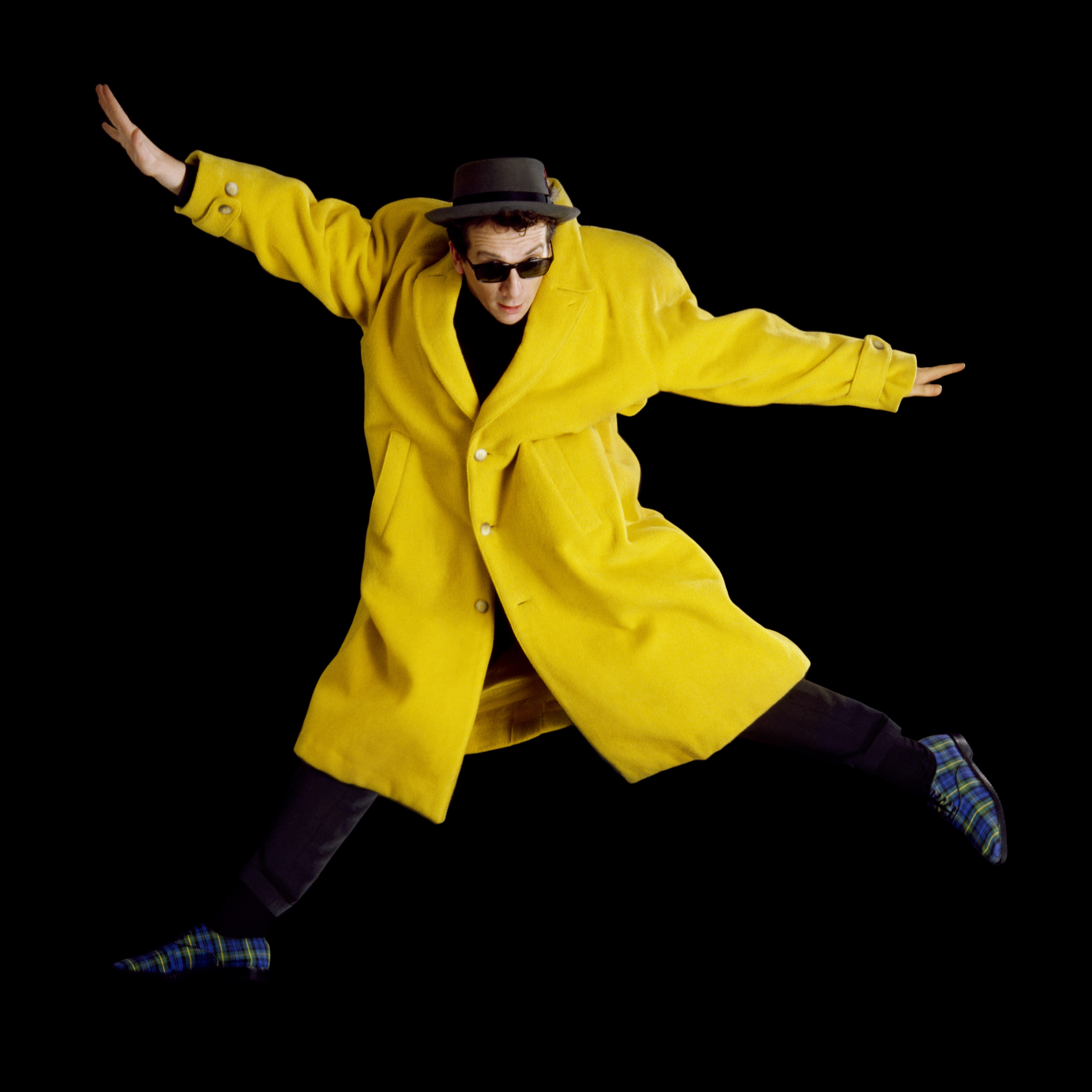 Elvis Costello Jumping 1986 If there is one thing Elvis Costello has in spades it's enthusiasm. No matter what it is he's doing he always puts his heart and soul into it. Around the time of King of America we did a studio session together, all went pretty normally, we shot close portraits, half lengths, photos with a guitar, all the usual stuff. I told him we had enough good photos and could call it a day, but he replied that he had one more thing he wanted to try. He disappeared into the dressing room and came out a few minutes later in this bright yellow coat. On many people it would look ridiculous, but Elvis has this ability to carry clothes well and it actually suited him. It did not seem to me that you could do anything other than full length shots in this outfit, he even had these black watch plaid shoes to top off the outfit. So I suggested he just jump up in the air a few times. He happily did this, doing his best Pete Townshend impersonations, but for the last couple of frames he flung his arms out wide creating the photo you see here. It appeared first in the Face, we always offered Nick Logan out best work and he ran this photo across a spread. To me it sums up EC perfectly.