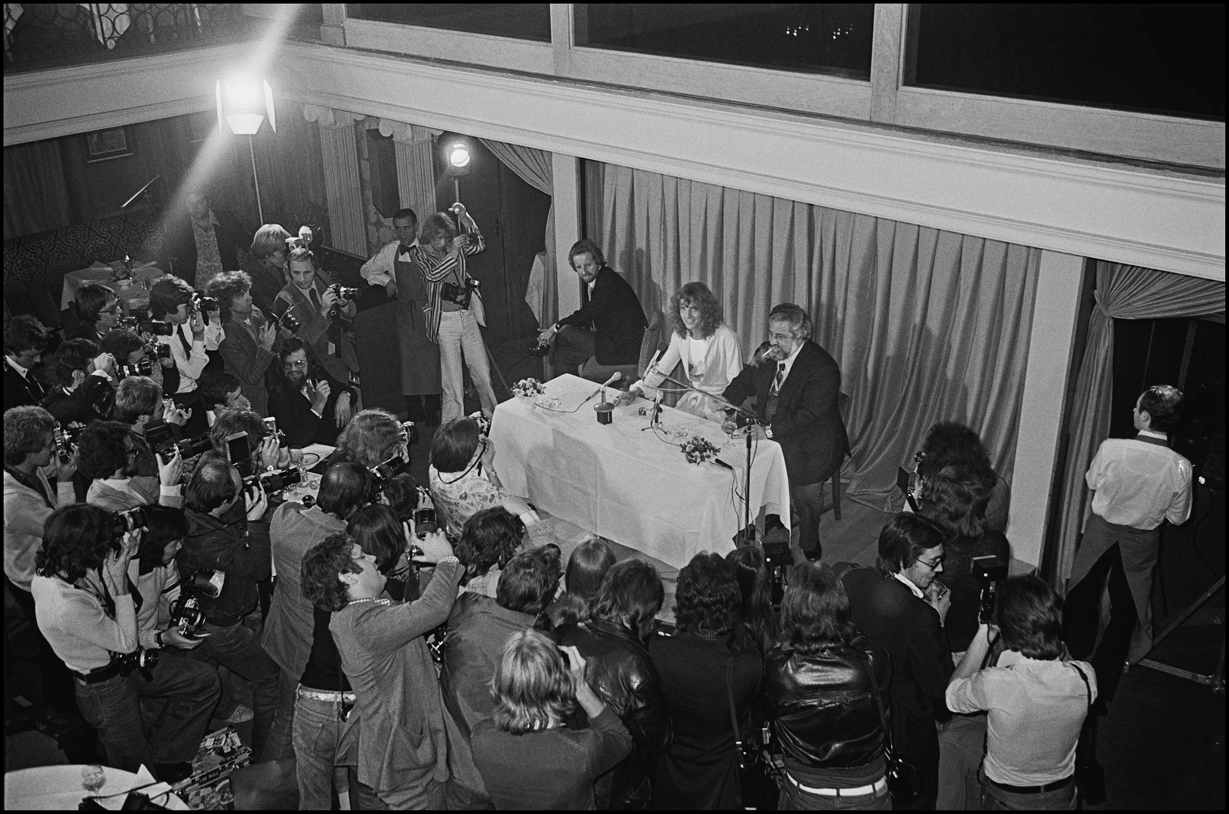 Peter Frampton's Press Conference - The Paparazzi Frampton Comes Alive, if ever there was phenomenon in the mid 70's then this was it. He was a cute blonde, formerly of Humble Pie, he took America by storm wearing a Baby Blue Satin Suit and by releasing a Live Double Album. Eventually of course he came back to the UK, and the chaos continued, his Photo Call attracted more photographers than I had ever seen assembled for a PopStar. It was a Zoo, there was no way of getting close, no chance of a decent picture, in fact I nearly went home thinking it was a pointless exercise. But, then I spotted a dark and dimly lit staircase, I ventured up it and found myself on a deserted balcony, the perfect place for pictures, of course he was small in the frame, and sitting next to his Manager, Dee Anthony, he certainly looked small. I got my picture, it was pretty good too, there he was, surrounded by photographers, and looking straight at me. The paper loved it and ran it big. People wondered how I did it, how could I get him to look right at me. It was easy, I just shouted "Peter, Up Here" at the top of my voice. Job done...