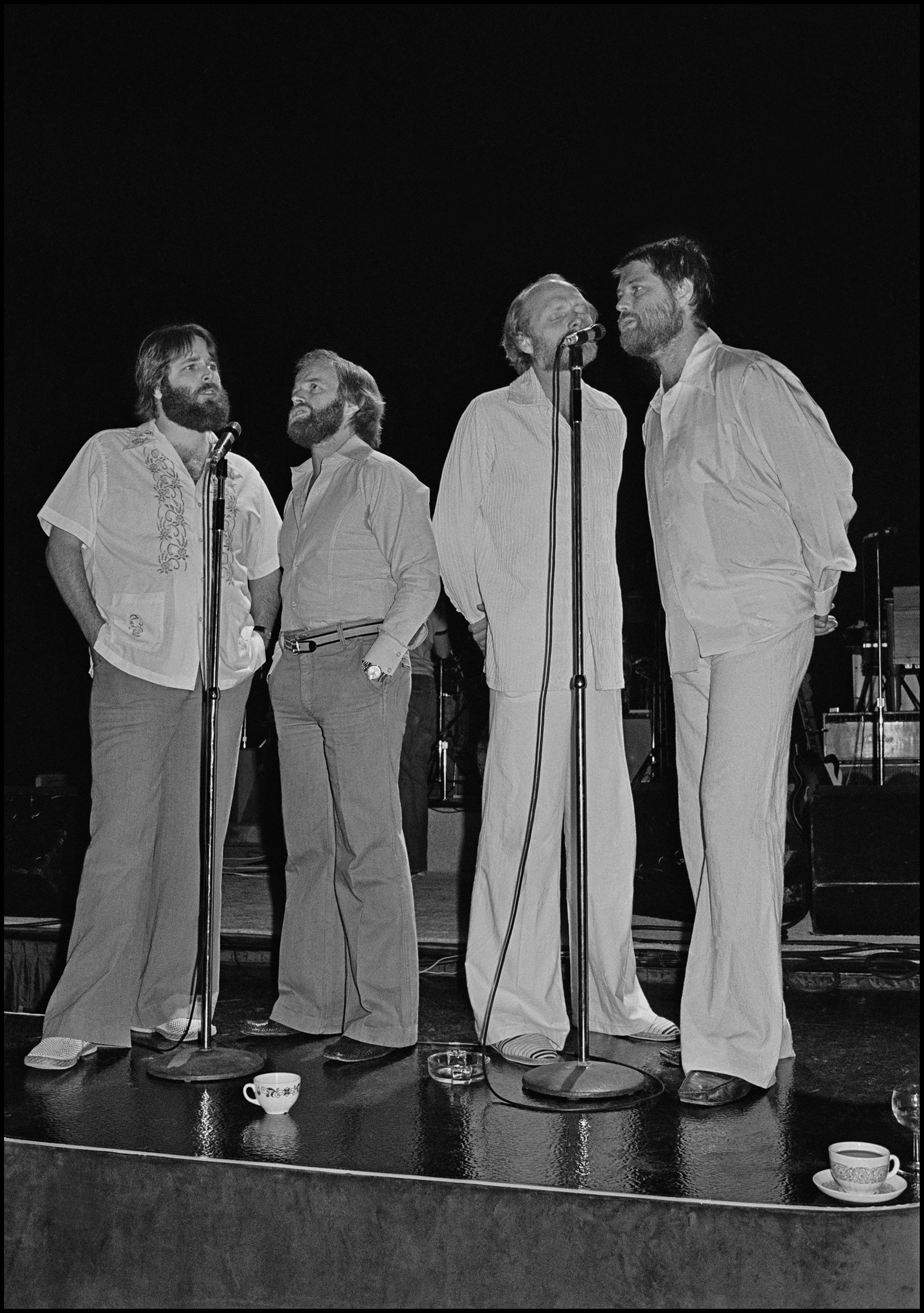 The Beach Boys - Dorchester Hotel 1977 You could describe my life as pure luck, not in getting the picture each time, that part was relatively easy, but just in terms of who I got to see and the circumstances under which saw them. This photo definitely comes into that category. Back in the 70's the Record Companies were flush with cash and were happy spending it on themselves. CBS took this to the extreme with their Annual Conventions, these multi day events consisted of flying hundreds of Record Co Execs to some exotic location and then have their best Artists perform for them, Bruce Springsteen played the 1975 one in New Orleans. In 1977 they had it in London, on the morning of the first day Elvis Costello started busking on the street outside the Grosvenor Park Hotel, his hope was to get the attention of the CBS people, instead he became a person of interest to the Police and got arrested. Luckily he got out of jail in time to play Dingwalls later that night. The highlight of the Convention was a performance by the Beach Boys on the final night. I was pretty good at sneaking into places unnoticed and so, along with my friend Kevin, we slipped into the Ballroom via the service entrance in mid afternoon. I did not know if we would see anything but it was worth a try. We were hiding behind piles of stacked chairs when the place started to get quite active. Then, in front of out star struck eyes, out came the Beach Boys to do a Soundcheck. Better yet, Brian Wilson was with them, he had not bern seen in public or performed with them since his breakdown and in NME land this was scoop central. I stayed hidden for the first three numbers, behind Brian was his Doctor/Psychiatrist who would massage his shoulders between numbers, he also shouted out the words to the songs acting as a sort of full time prompter to the shell shocked looking Brian. Figuring that they might only do four songs I took my chance and darted out from my hiding place, I scurried to the front of the stage and took five very quick frames before running out of the Ballroom, thru the Hotel Lobby and out on to Park Lane. I had my photo and nobody was going to take it away from me. After developing it I noticed the two teacups on the floor in front of their feet. To me it really adds to the impact of the image by adding just a hint of the surreal to it. As I said I was really fucking lucky, I really did have the best job in the world...